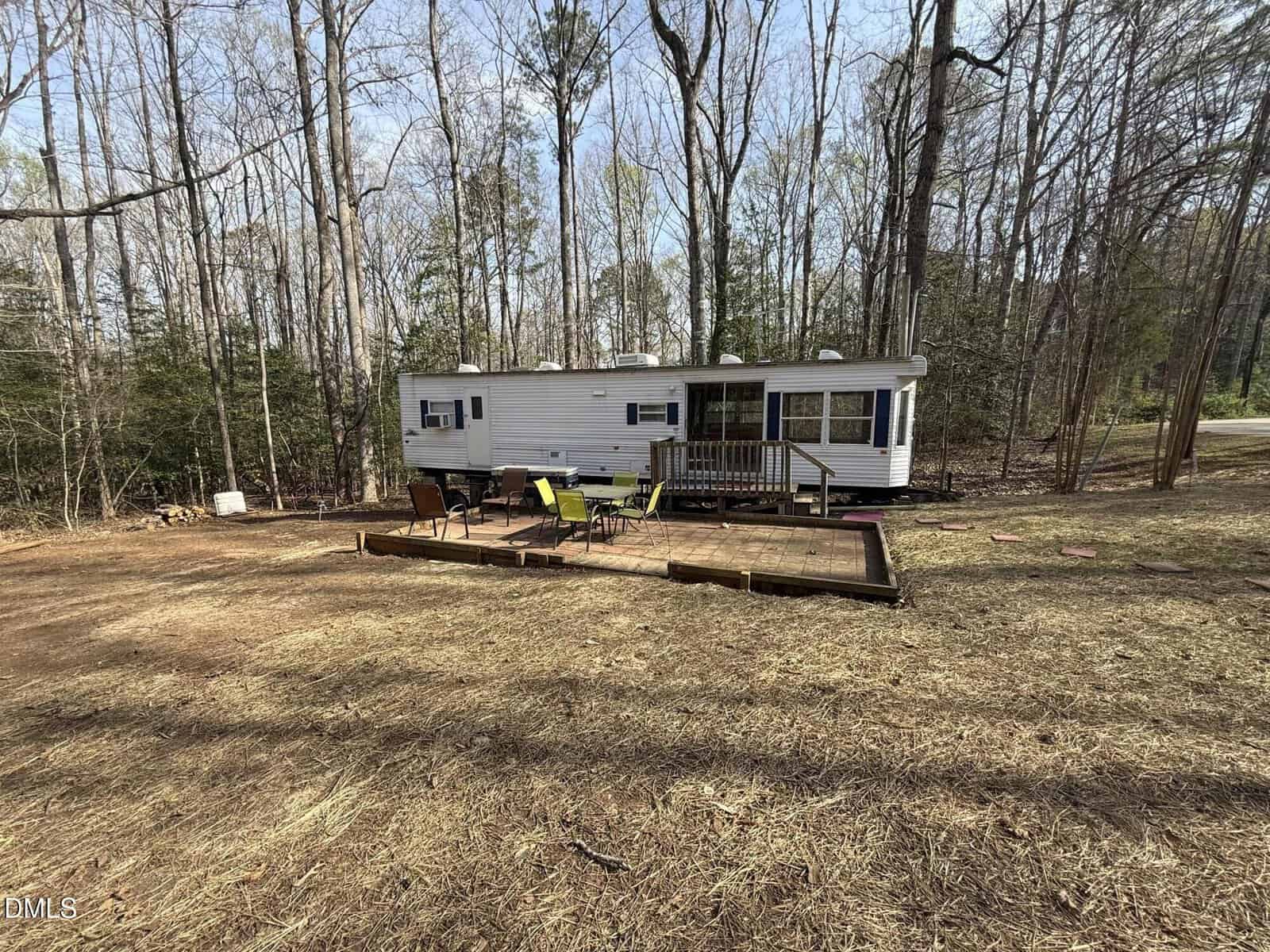 A white mobile home with a small covered porch is situated in a clearing surrounded by tall trees. In front, there is a wooden deck with yellow and black outdoor chairs and a table. The ground is mostly dirt and dry grass.