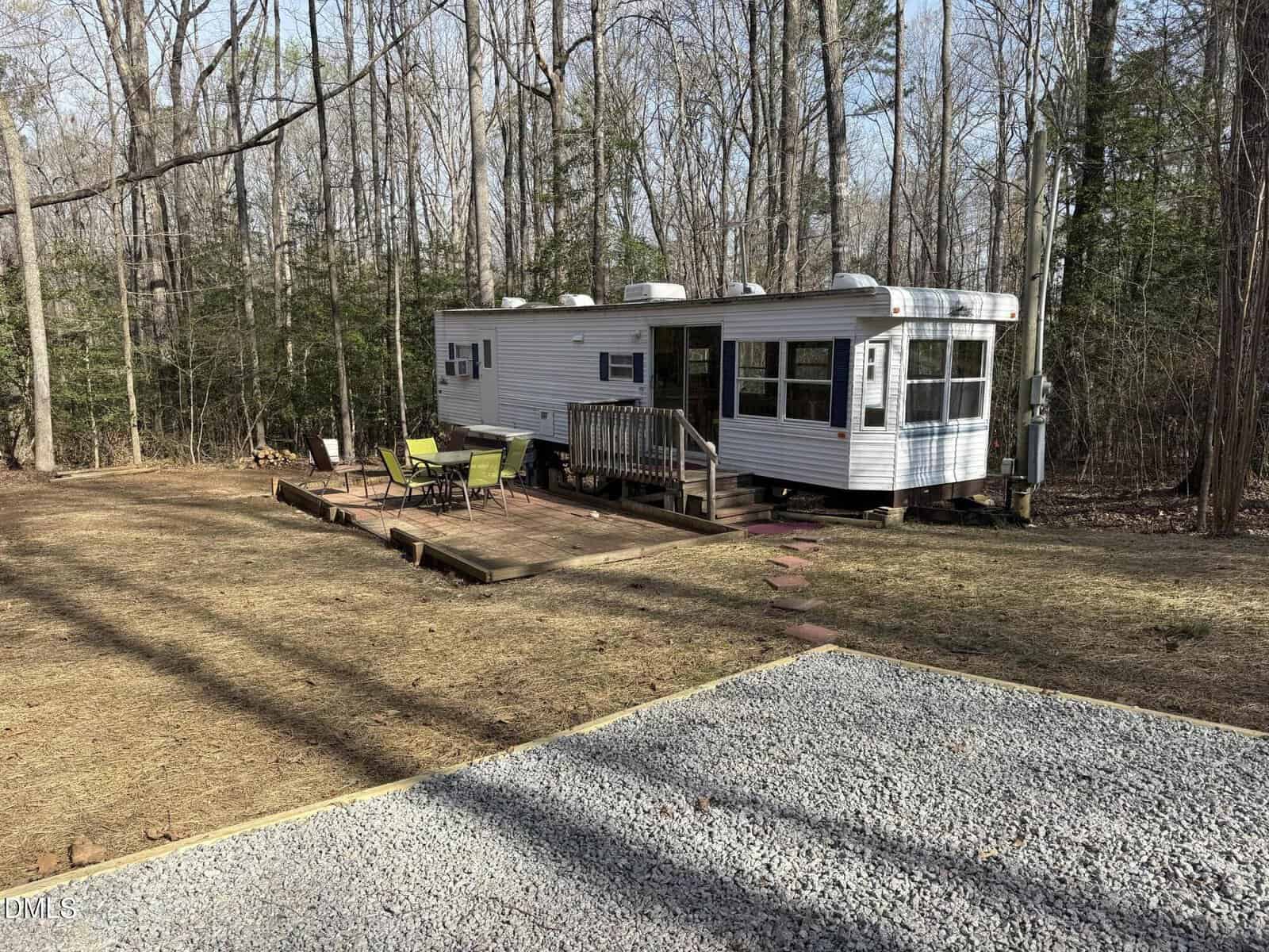 A white mobile home sits in a wooded area with a wooden deck. On the deck are a table and green chairs. The ground is mostly bare with some grass, and a gravel area is visible in the foreground.