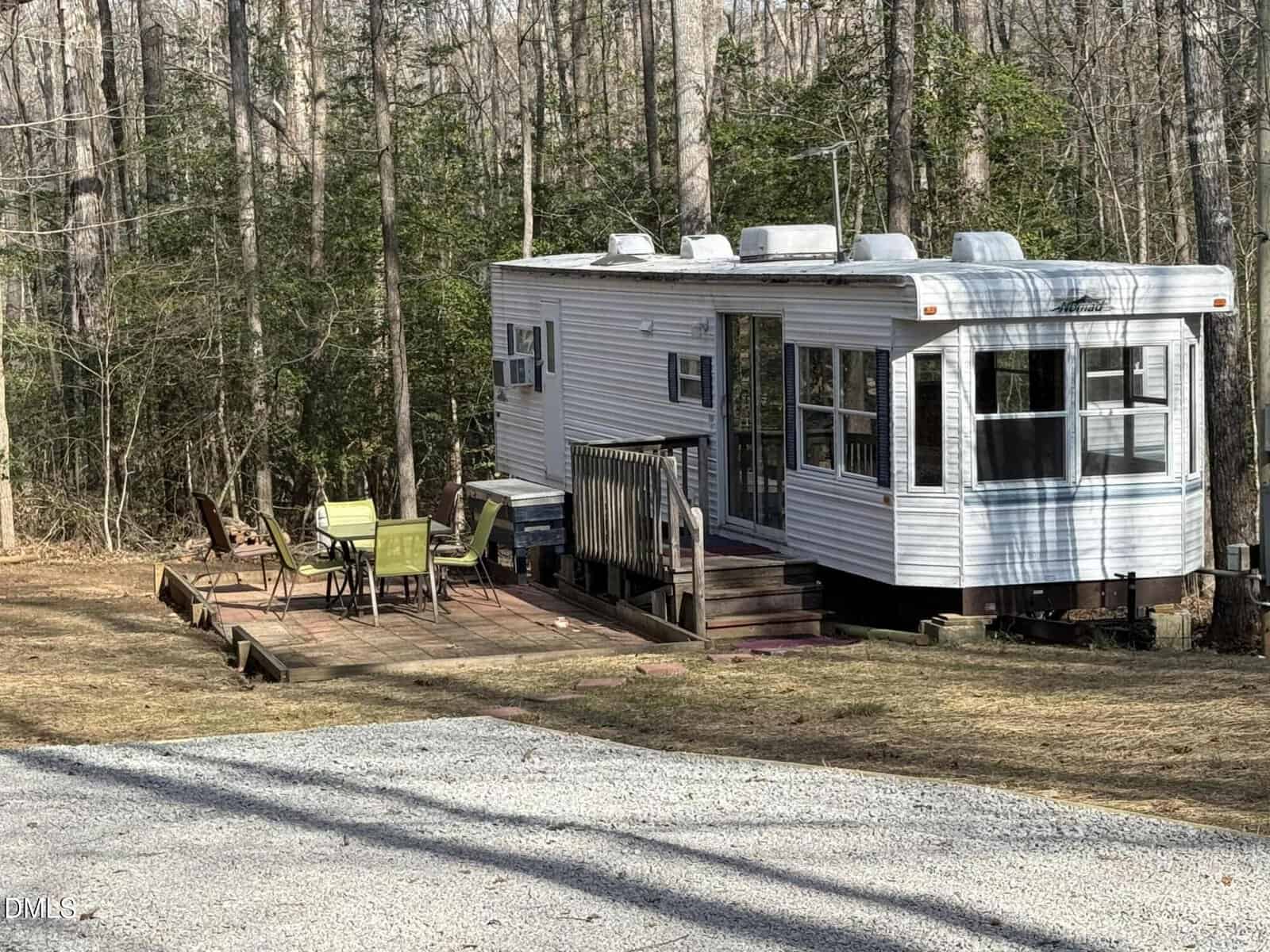 A small white mobile home with several windows is situated in a wooded area. A wooden deck with patio chairs and a table is attached to the home, and a gravel driveway is visible in the foreground.