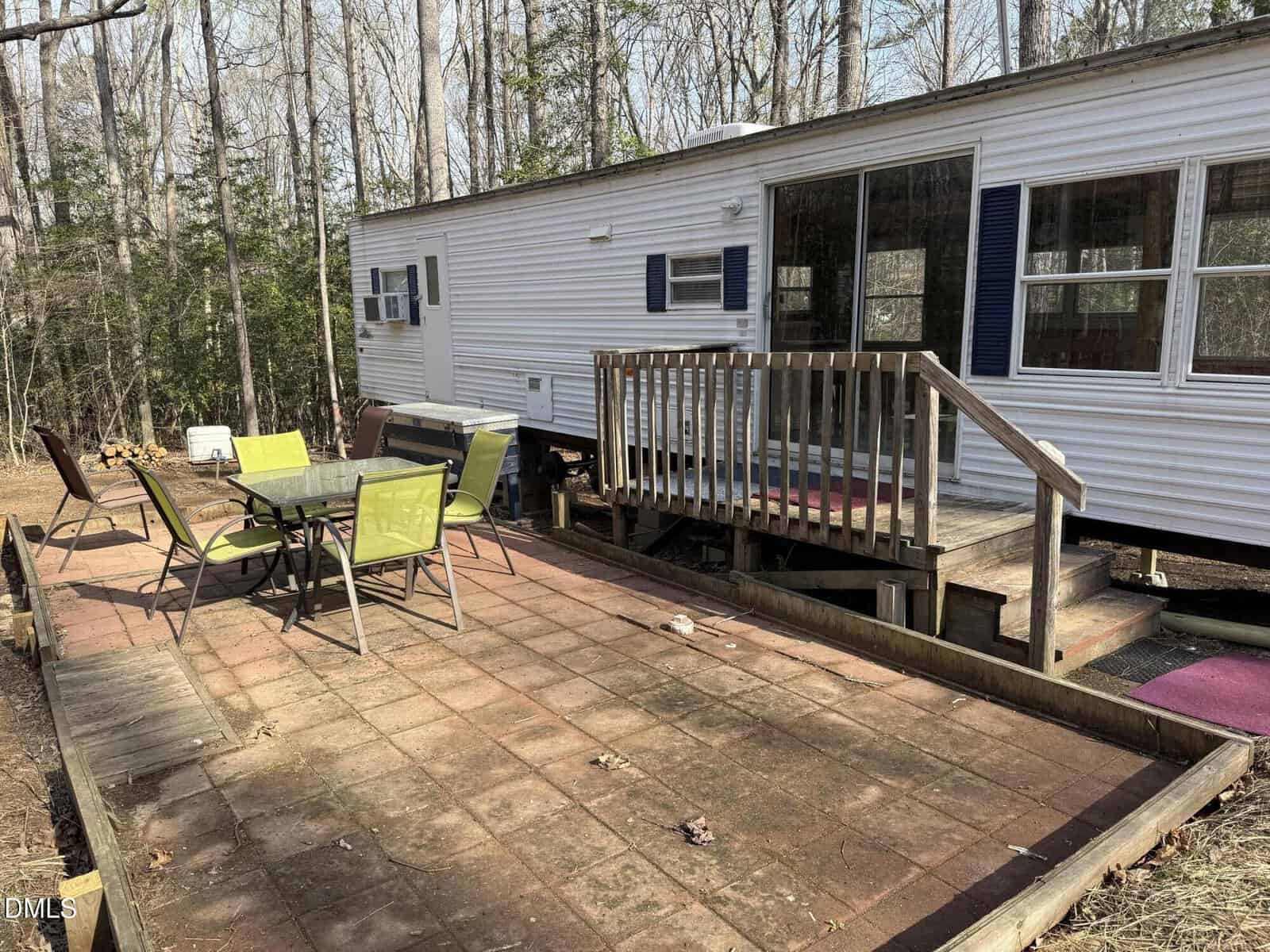A white trailer with a small wooden porch is shown. In front, there is a patio with four green and beige chairs around a glass table. The area is surrounded by trees and leaf-covered ground.