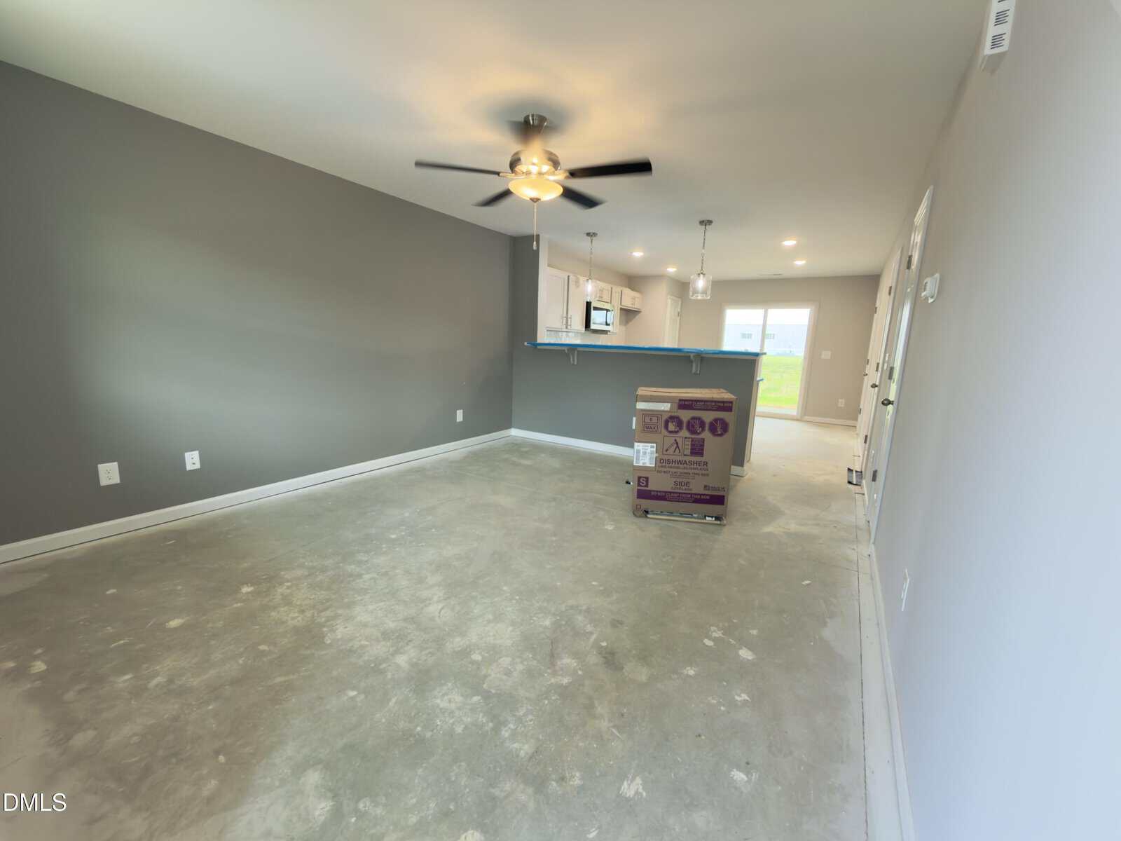 Unfurnished living room with concrete floors, gray walls, ceiling fan, and overhead light. Kitchen with white cabinets and appliances visible in the background. A large box is in the center of the room.