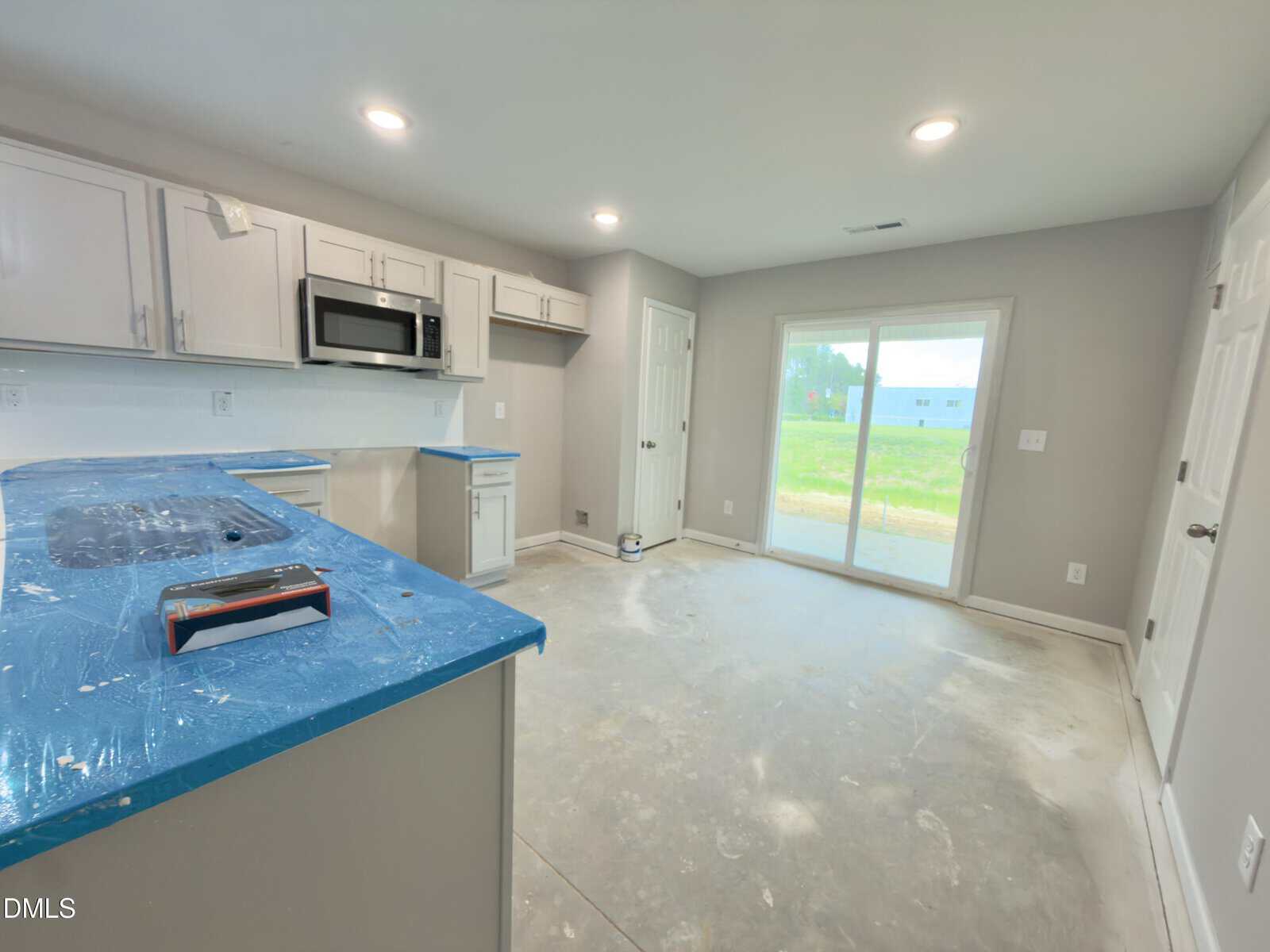 A kitchen with unfinished floors, covered countertops, white cabinets, a built-in microwave, and sliding glass doors leading to a backyard. The room appears to be under construction or renovation.