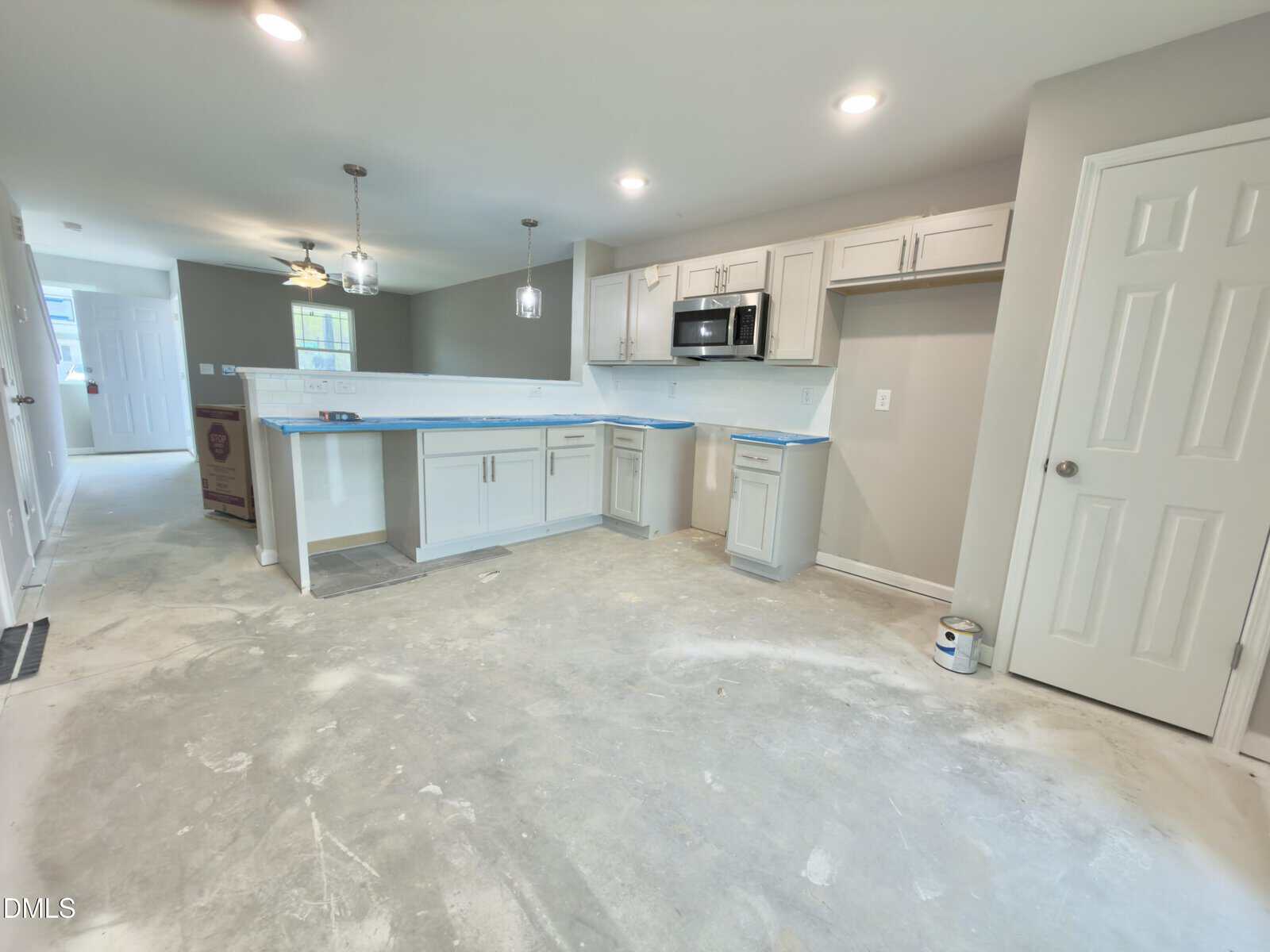 A kitchen under renovation with unfinished floors, white cabinets, a built-in microwave, and blue tape along the countertops. Paint supplies and protective coverings are visible. Walls are painted light gray.