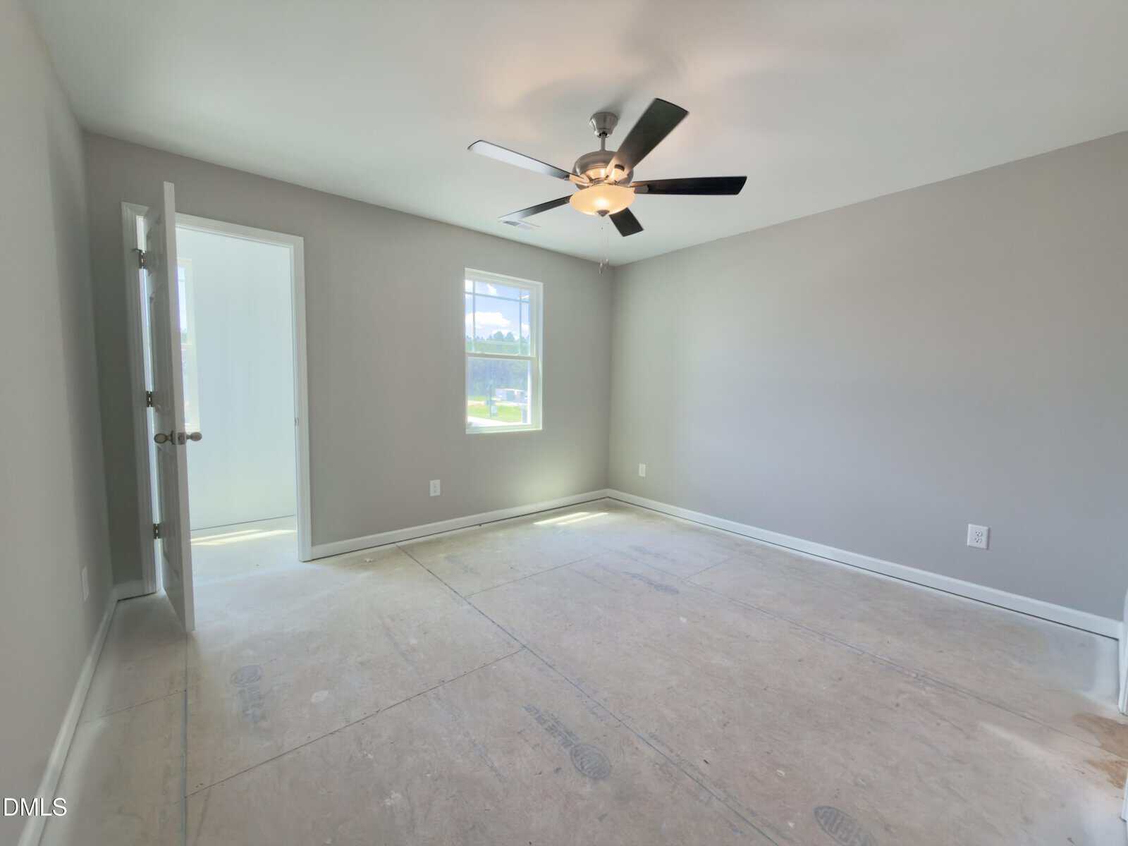 Empty bedroom with gray walls, a ceiling fan with light, large window, and open door leading to a bright bathroom. The floor is unfinished, showing protective covering.