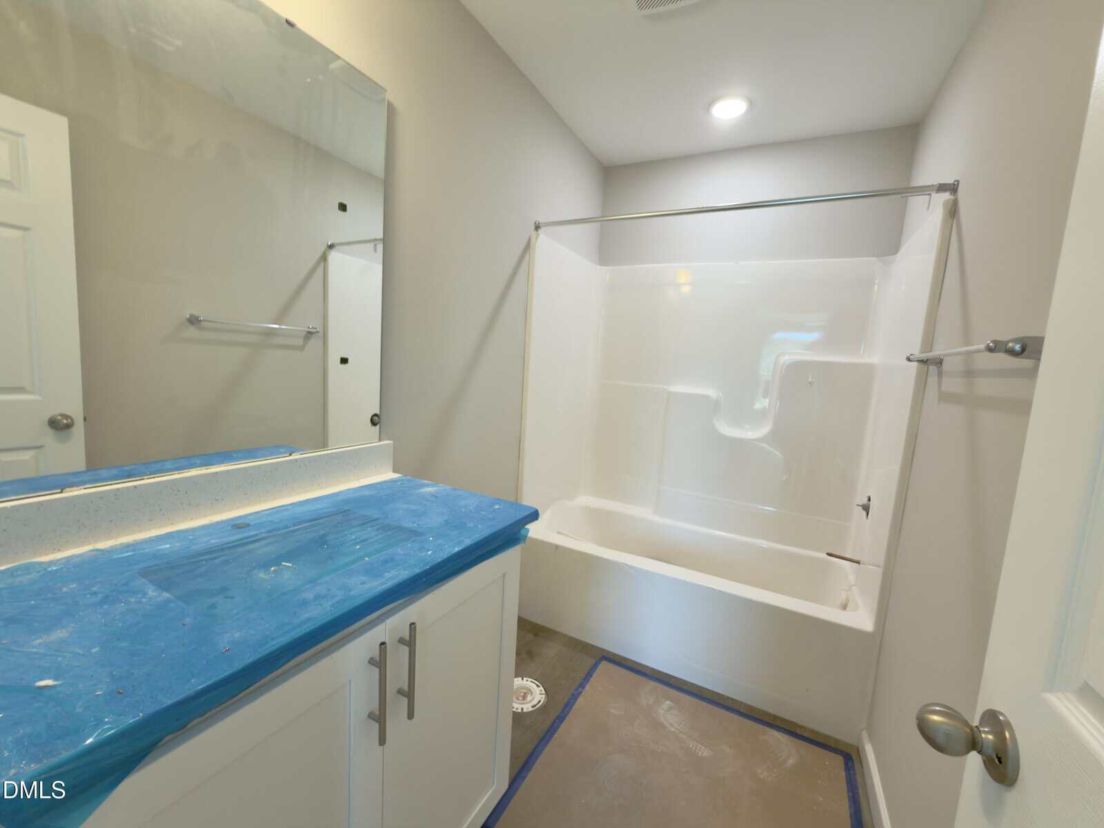 A bathroom under renovation with a covered blue countertop, white cabinets, a large mirror, a white bathtub with a shower, and unfinished flooring. Walls are painted light beige. A towel rack is installed next to the sink.
