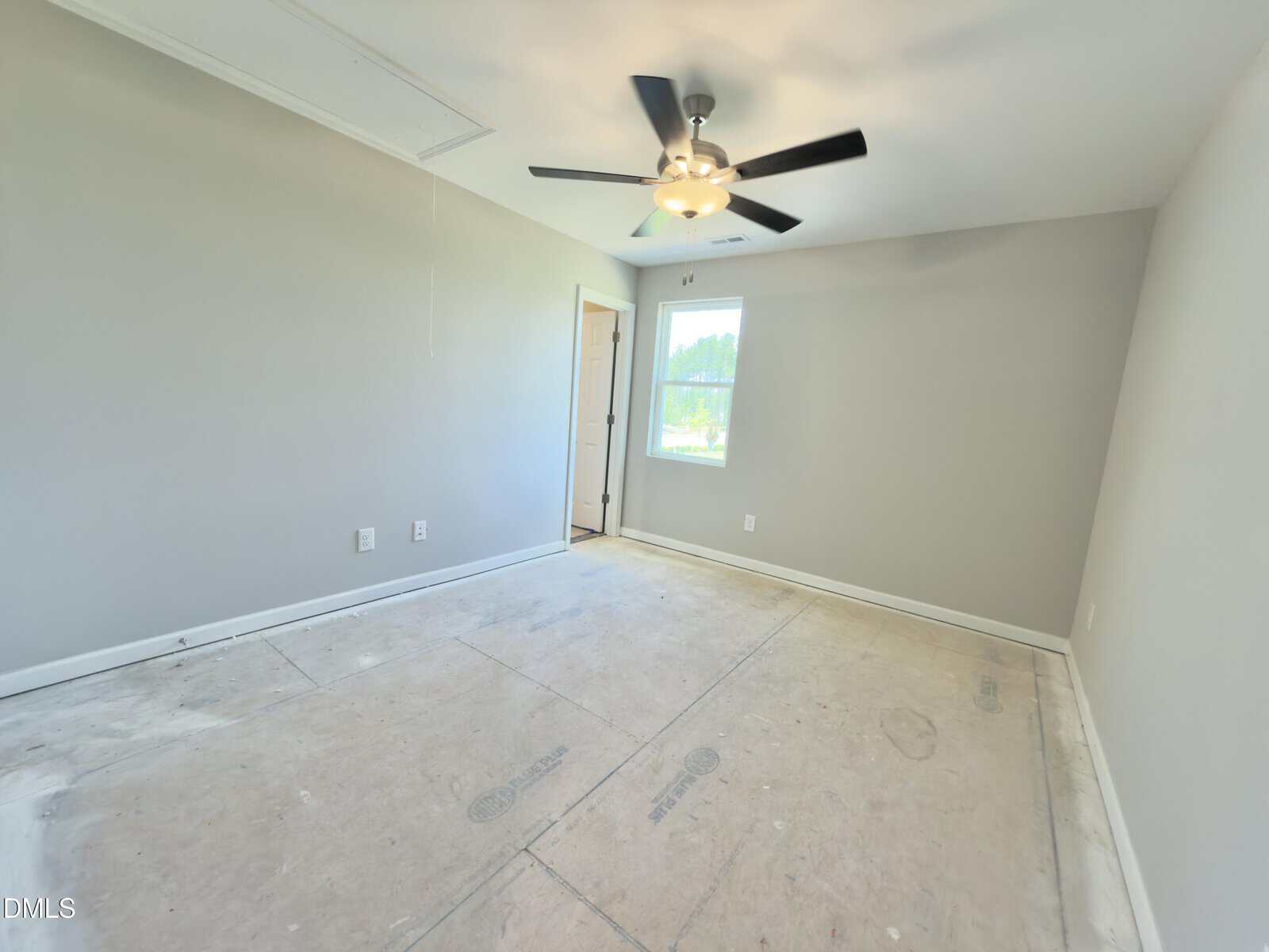 Empty room with light gray walls, a ceiling fan with light, a small window letting in natural light, and an unfinished floor. There is a closed door and visible electrical outlets on the wall.