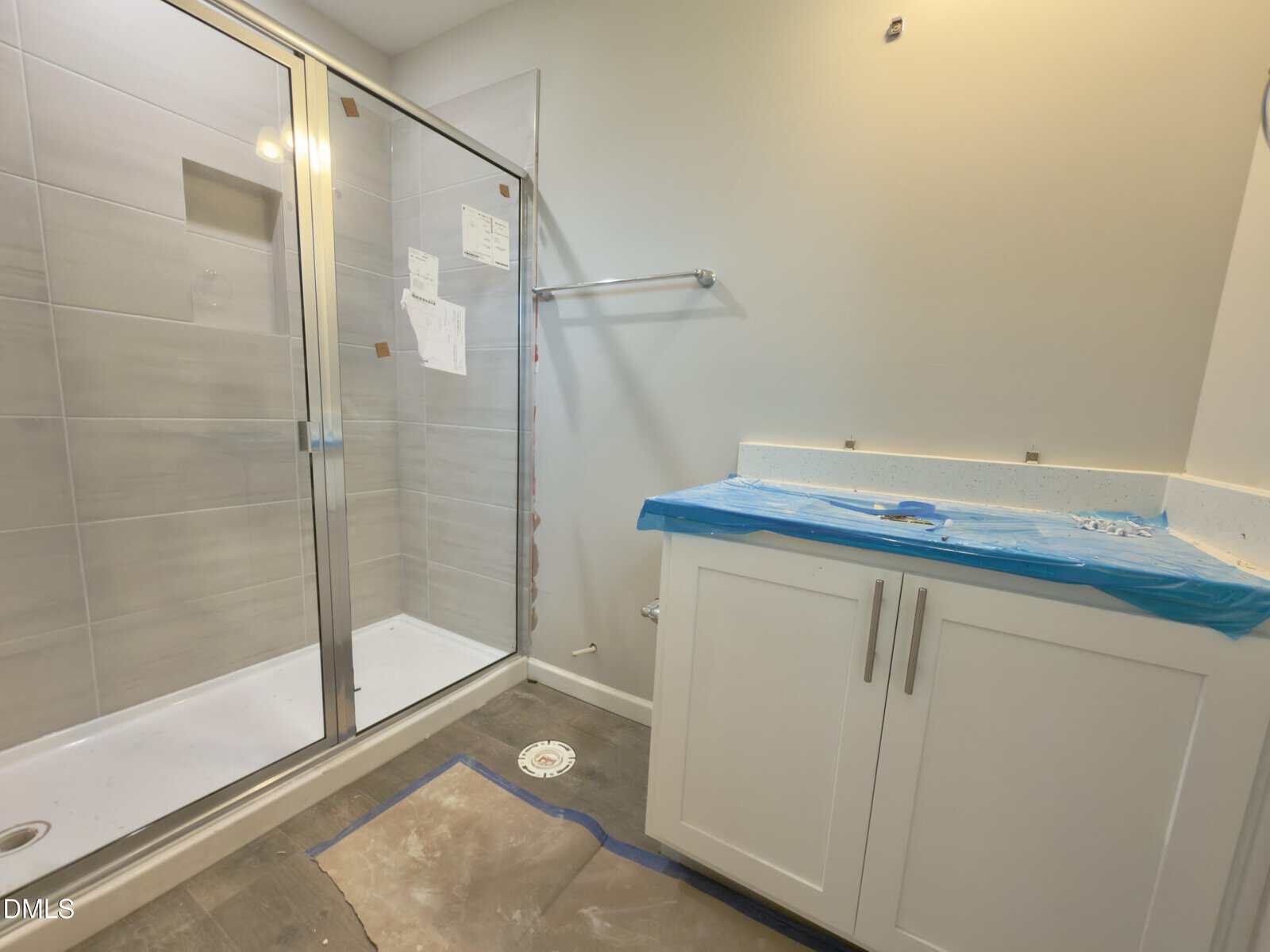 A bathroom under construction with a glass shower, gray tile, white vanity with blue protective covering, and unfinished floor and walls.