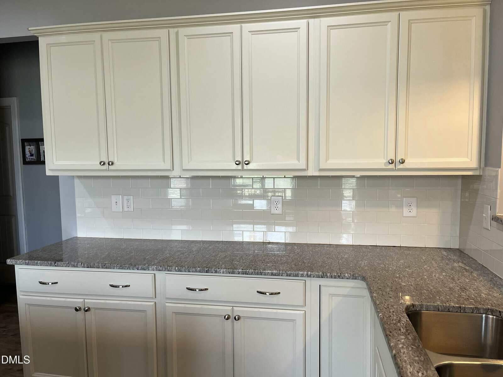 A kitchen with off-white cabinets, silver handles, a gray speckled countertop, a white subway tile backsplash, multiple electrical outlets, and a stainless steel sink in the bottom right corner.