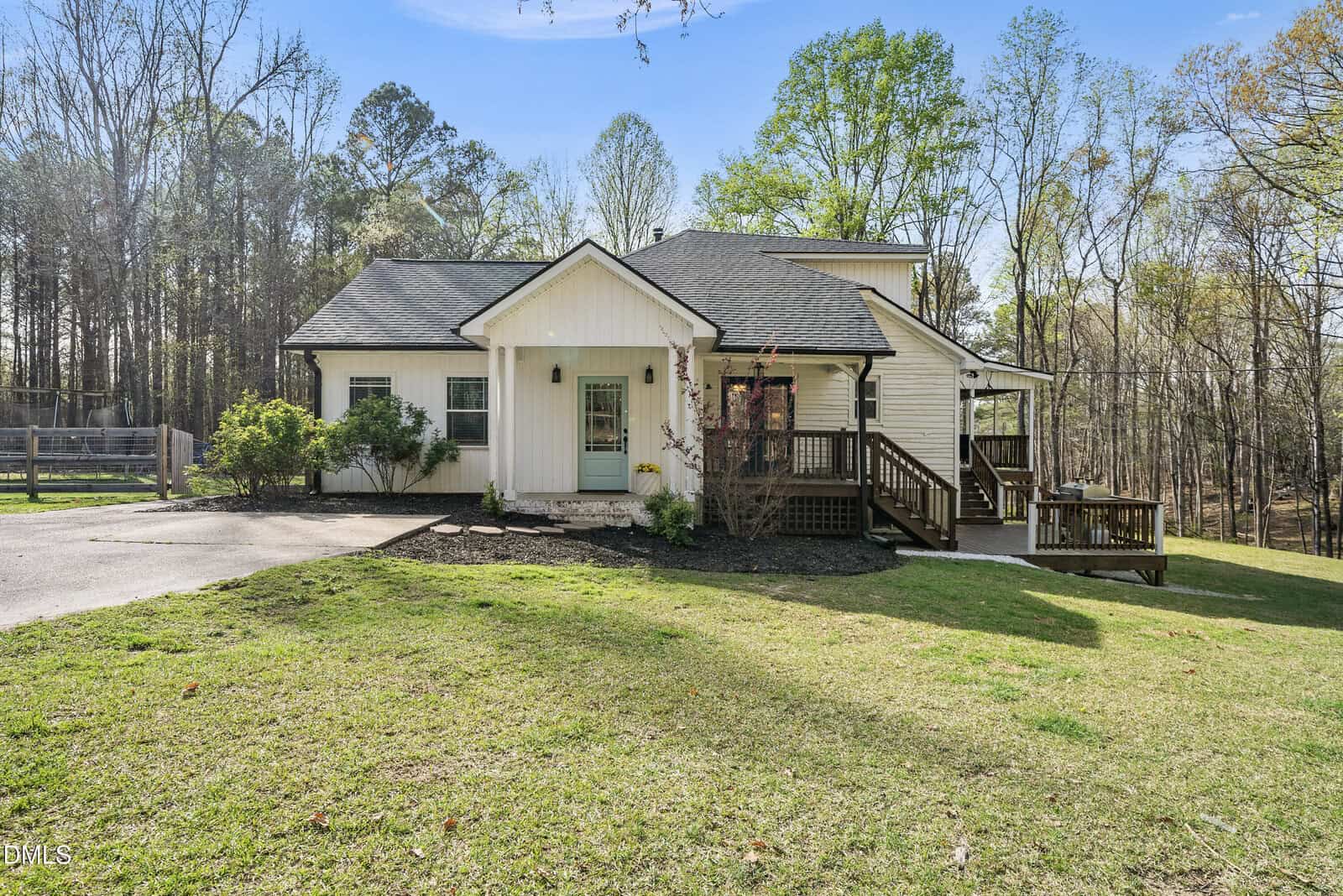 A modern farmhouse with white siding, a dark roof, front porch, and wooden deck sits on a spacious grassy lawn bordered by trees and a driveway under a clear blue sky.