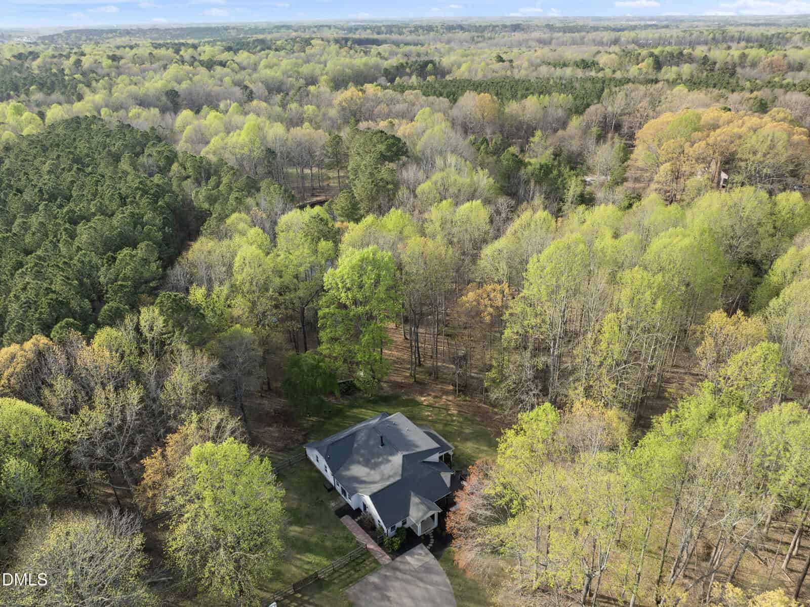 Aerial view of a gray house surrounded by a large expanse of forest with trees showing early spring foliage, under a partly cloudy sky. A driveway leads to the house, which sits in a clearing amid dense woods.