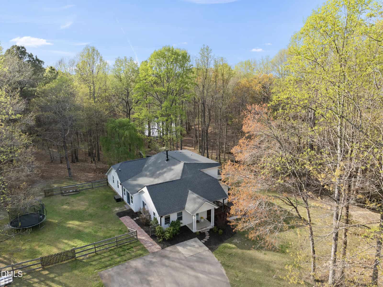 A white house with a dark gray roof sits among tall trees with spring foliage. A fenced yard with a trampoline is on the left, and a driveway leads to the front entrance. A pond is visible through the trees in the background.