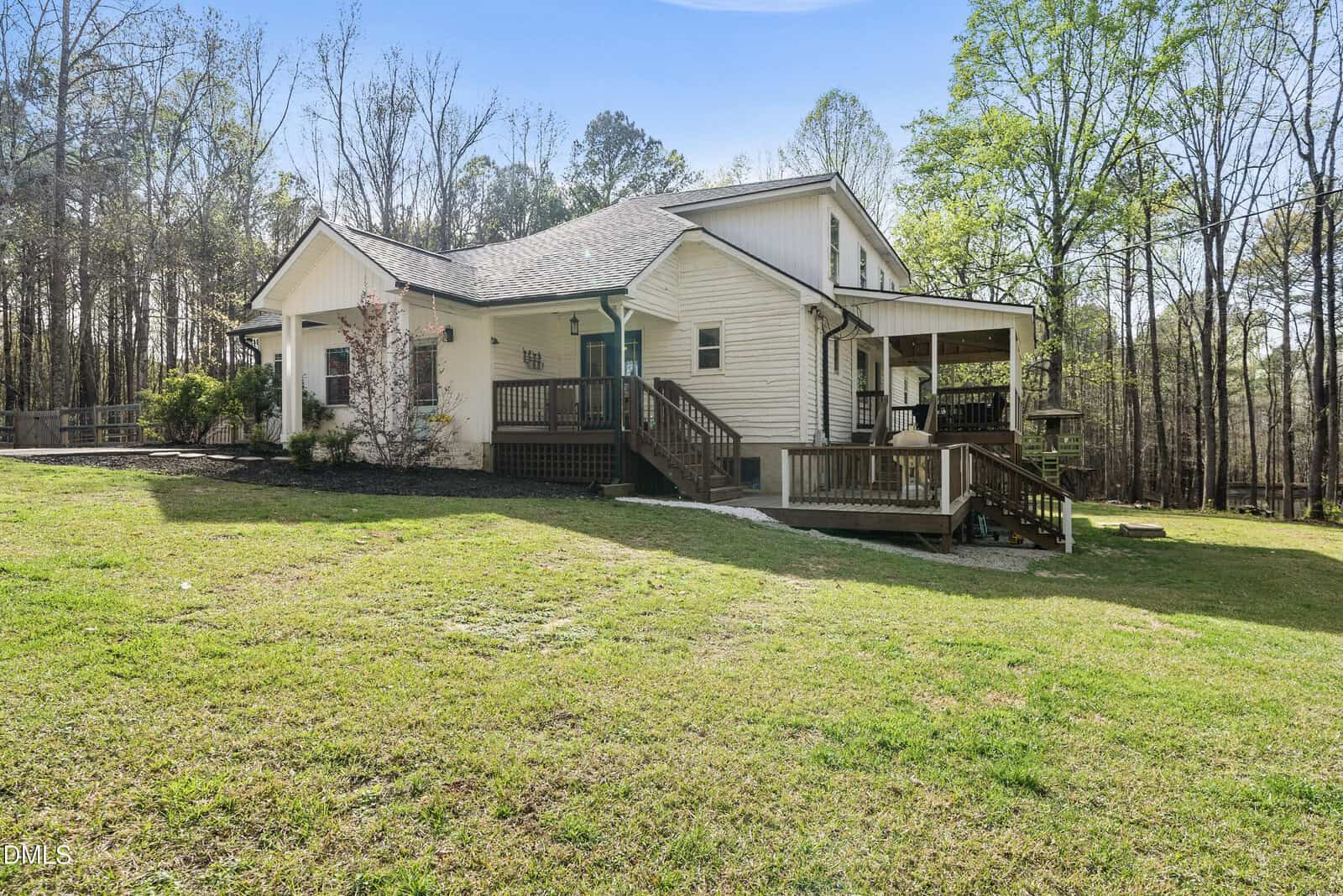 A white single-story house with a covered porch and wooden ramp, set on a spacious grassy yard with trees in the background under a blue sky.