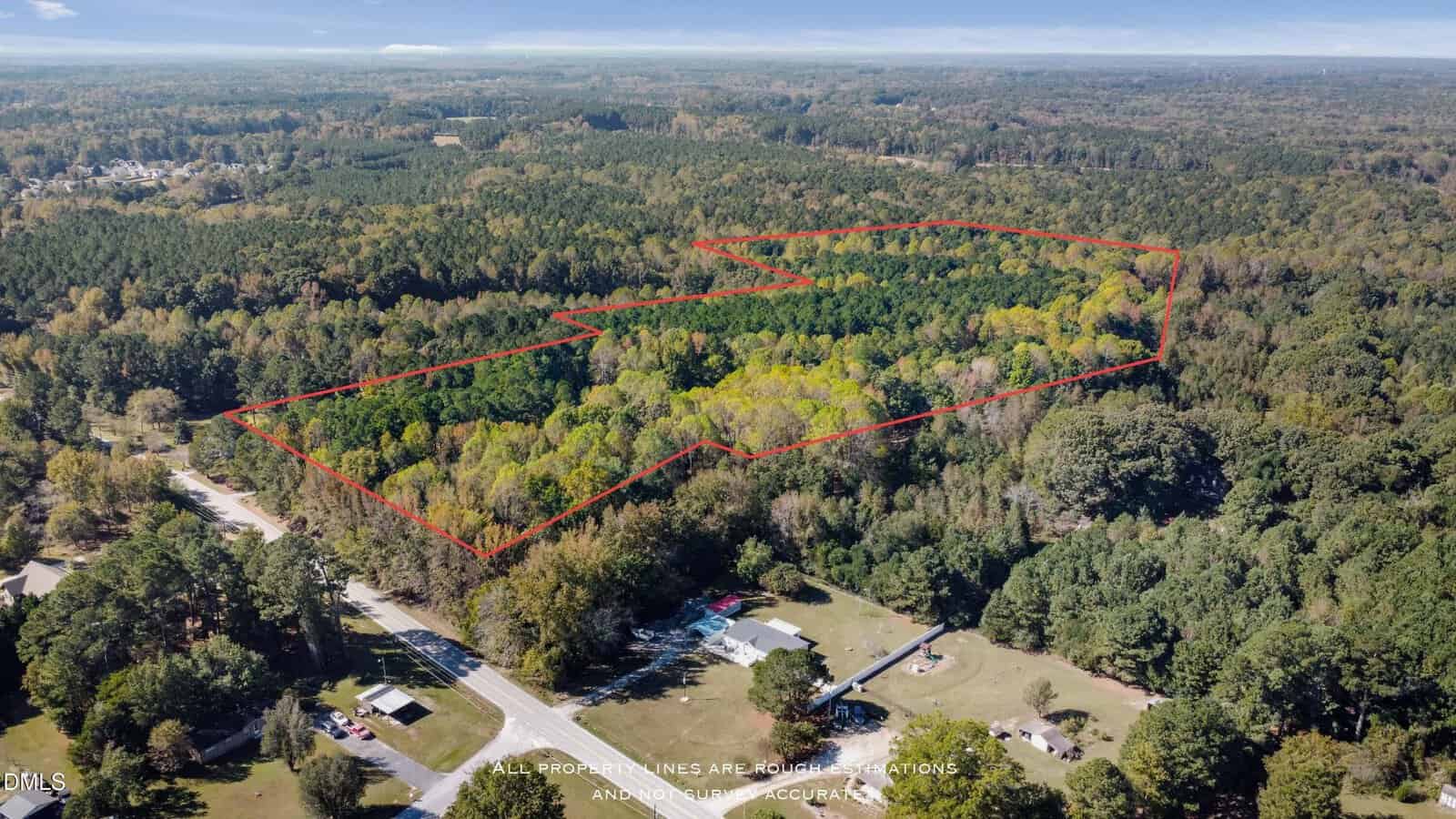 Aerial view of a forested area with a red outline marking property boundaries; nearby are some houses, a road, and more wooded land under a partly cloudy sky.