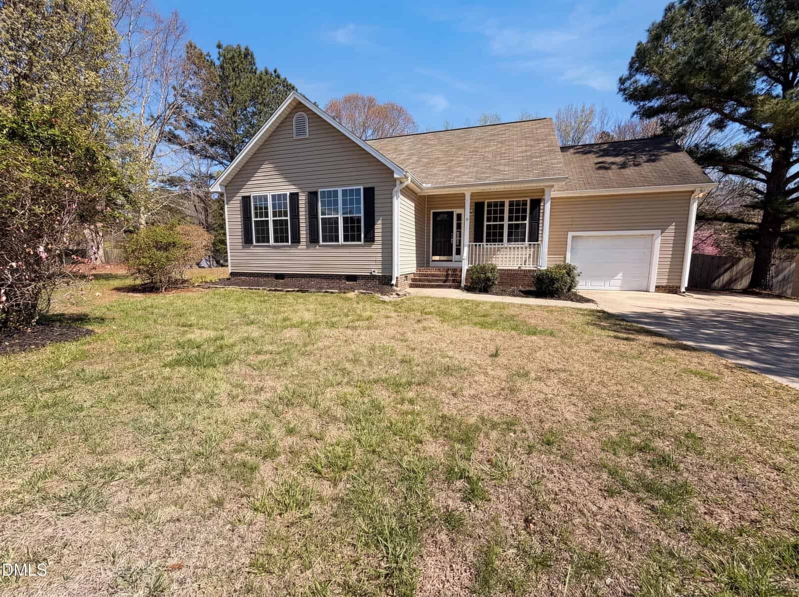 Single-story beige house with black shutters, a front porch, and an attached garage. The yard has patchy grass and some shrubs. A paved driveway leads to the garage. Trees surround the property under a clear blue sky.