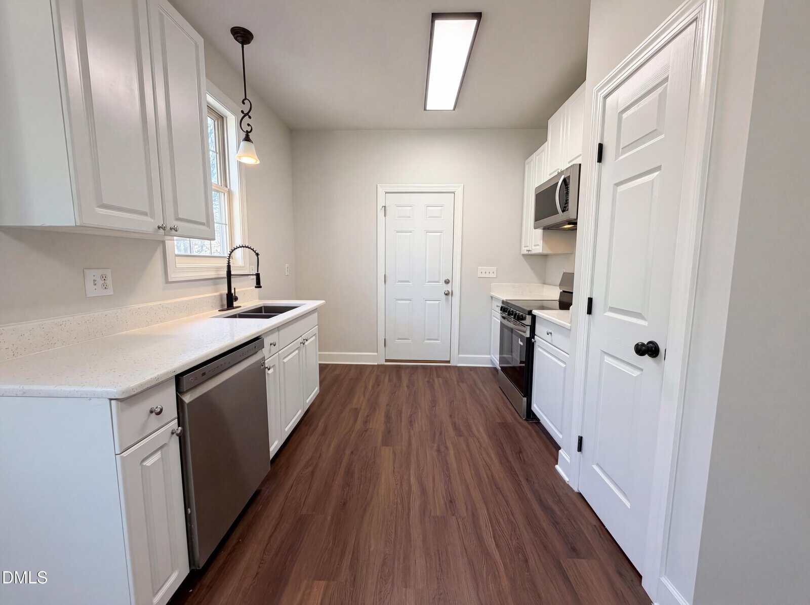 A modern kitchen with white cabinets, stainless steel appliances, wood-look flooring, and neutral walls. There is a window above the sink, a hanging light fixture, and a door at the end of the room.