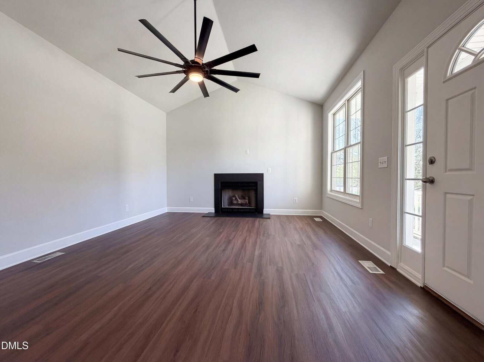 A bright, empty living room with vaulted ceiling, large ceiling fan, fireplace centered on the far wall, wood-look flooring, and a door with windows next to tall, double-pane windows.