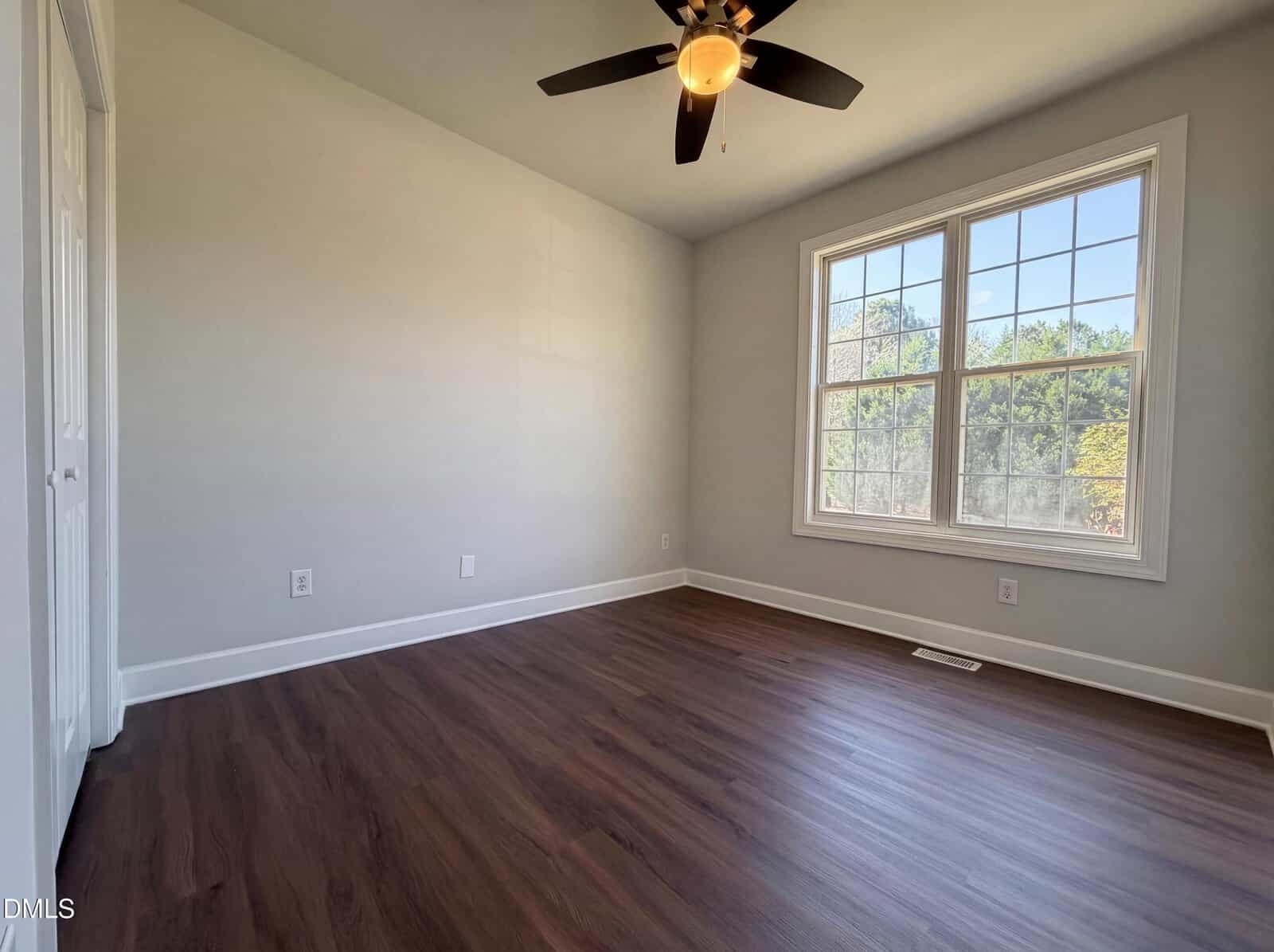 Empty room with light gray walls, dark wood flooring, a ceiling fan with a light, and a large double window letting in natural light. The room has a closet door and white trim.