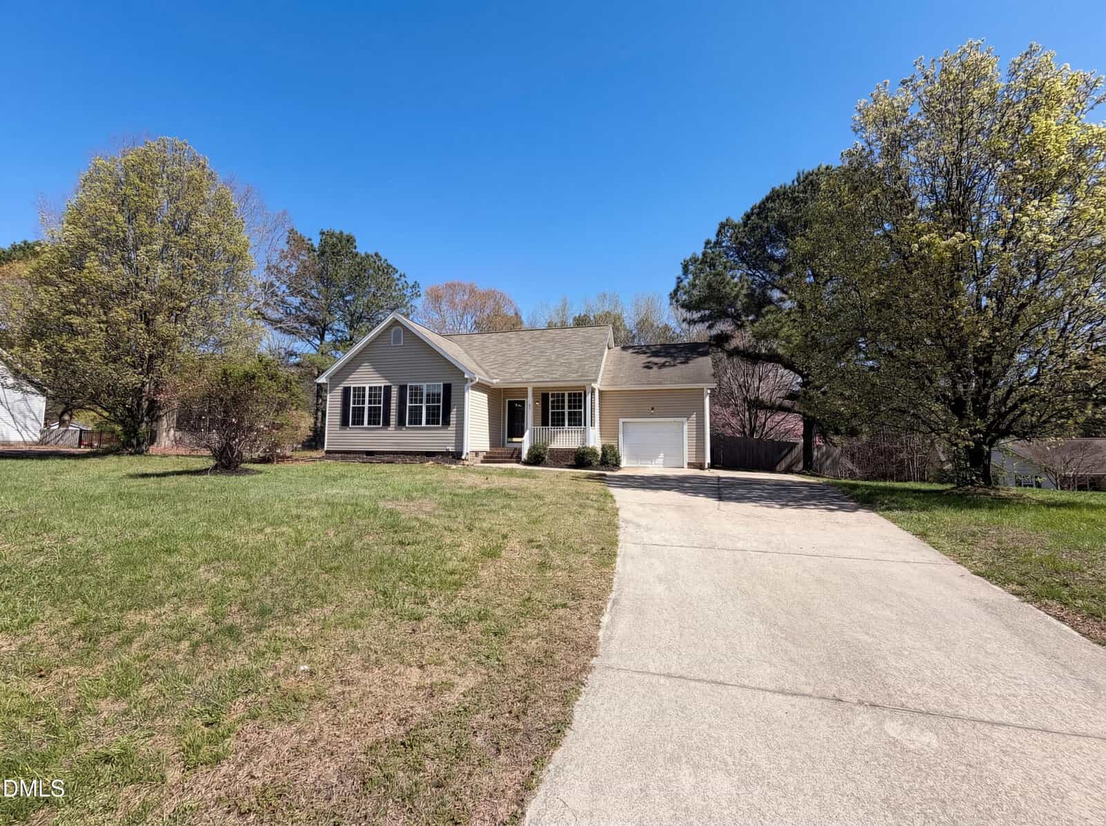 Single-story beige house with a front porch, attached garage, and large driveway. Surrounded by green grass, trees, and blue sky on a sunny day.