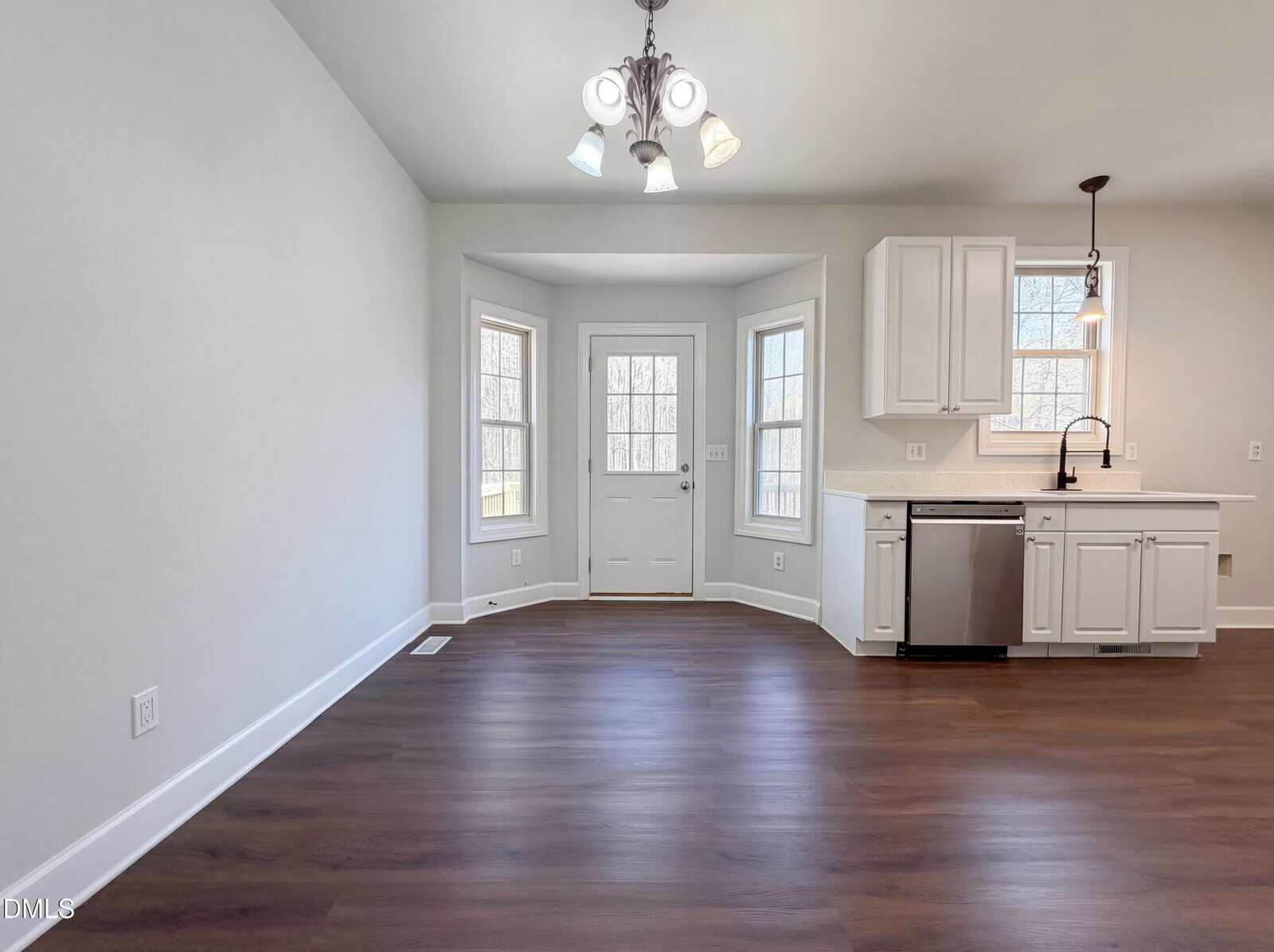 Bright kitchen area with white cabinets, stainless steel dishwasher, dark faucet, wood flooring, a bay window, and a door leading outside. Overhead lighting fixture is visible. Walls are painted light gray.
