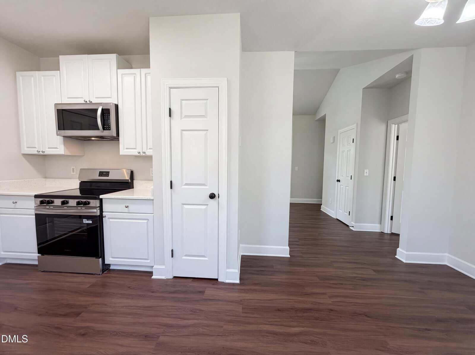 Modern kitchen with white cabinets, stainless steel appliances, and wood-style flooring; adjacent hallway with white doors and light gray walls visible in the background.