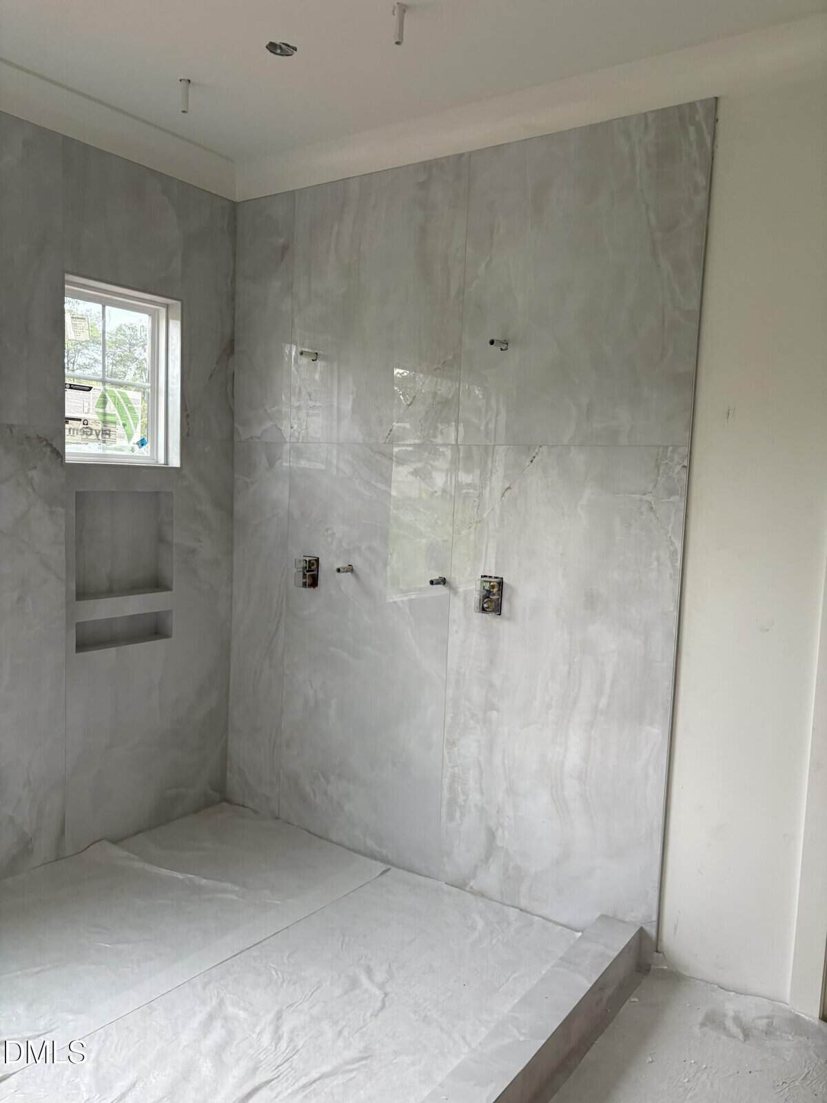 A modern, unfinished bathroom with large gray marble tiles, built-in wall niches, a small window, and exposed plumbing fixtures for a shower.