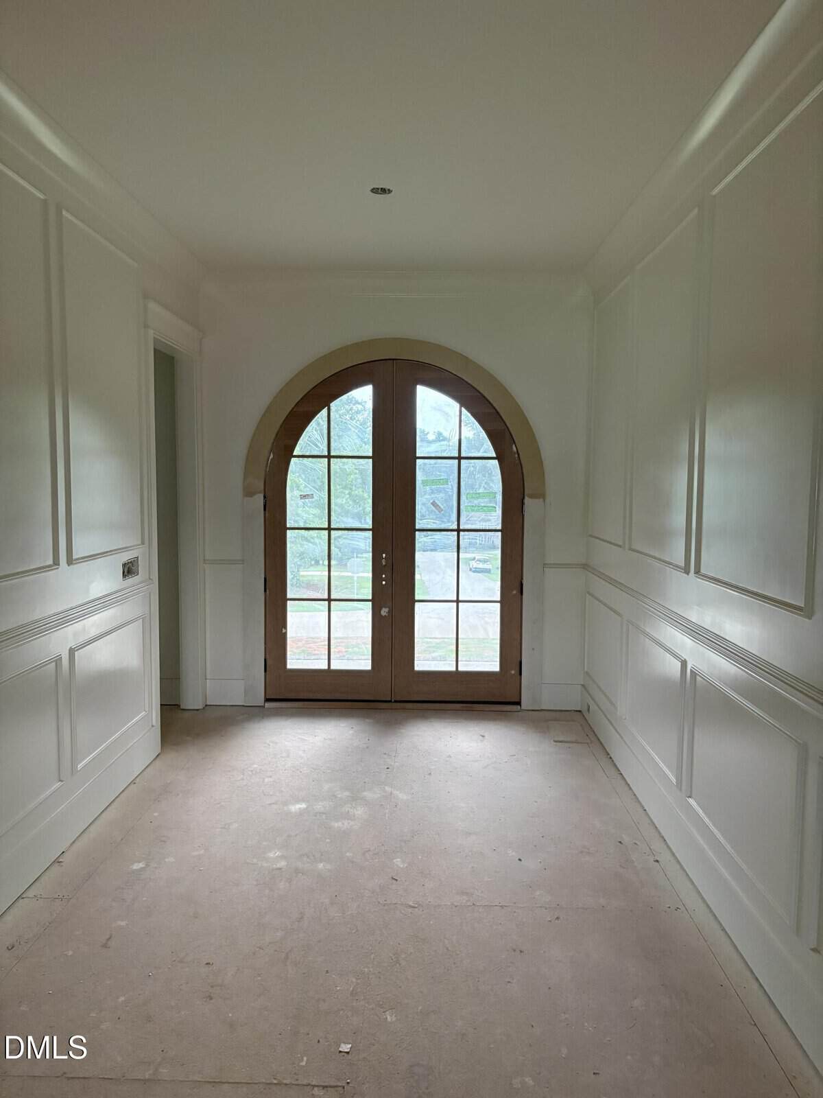 A hallway with white paneled walls and double wooden arched doors with glass panes, letting in natural light and revealing a view of greenery outside.