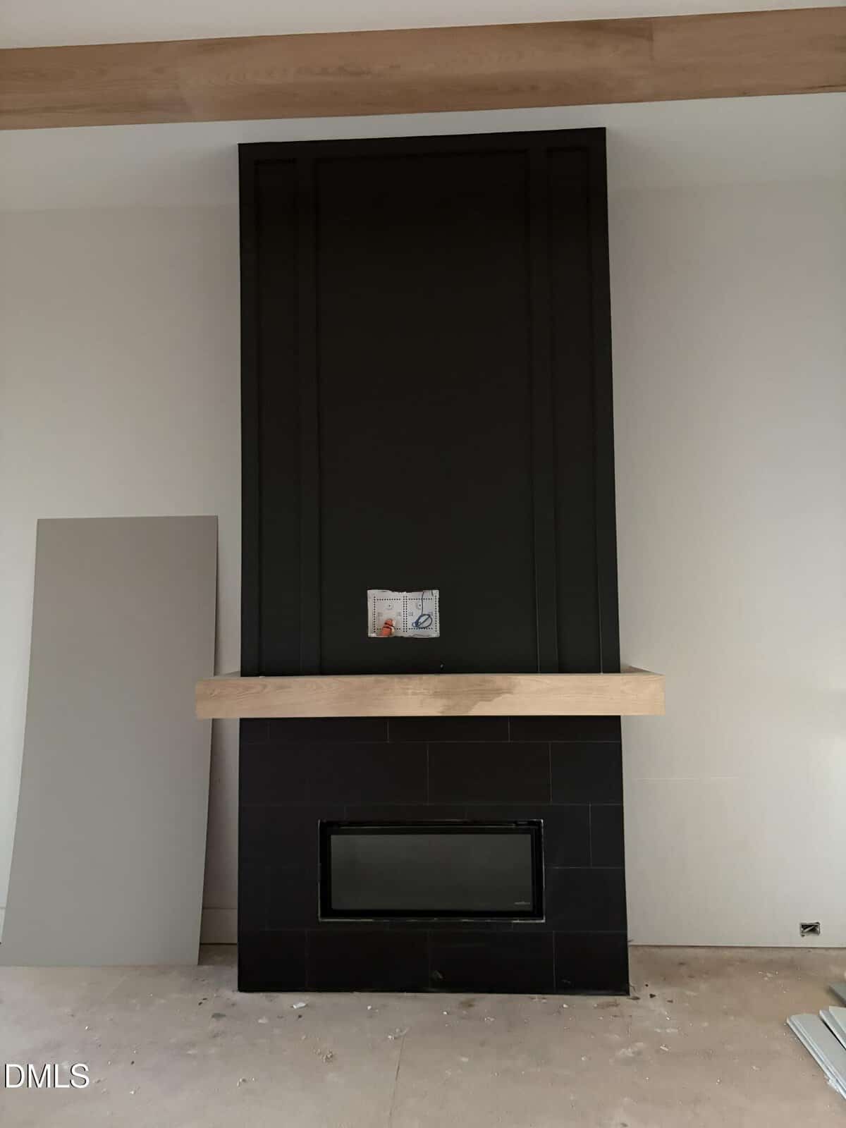A modern black fireplace with a wooden mantel, surrounded by light-colored walls. A panel leans against the wall on the left, and unfinished flooring is visible.