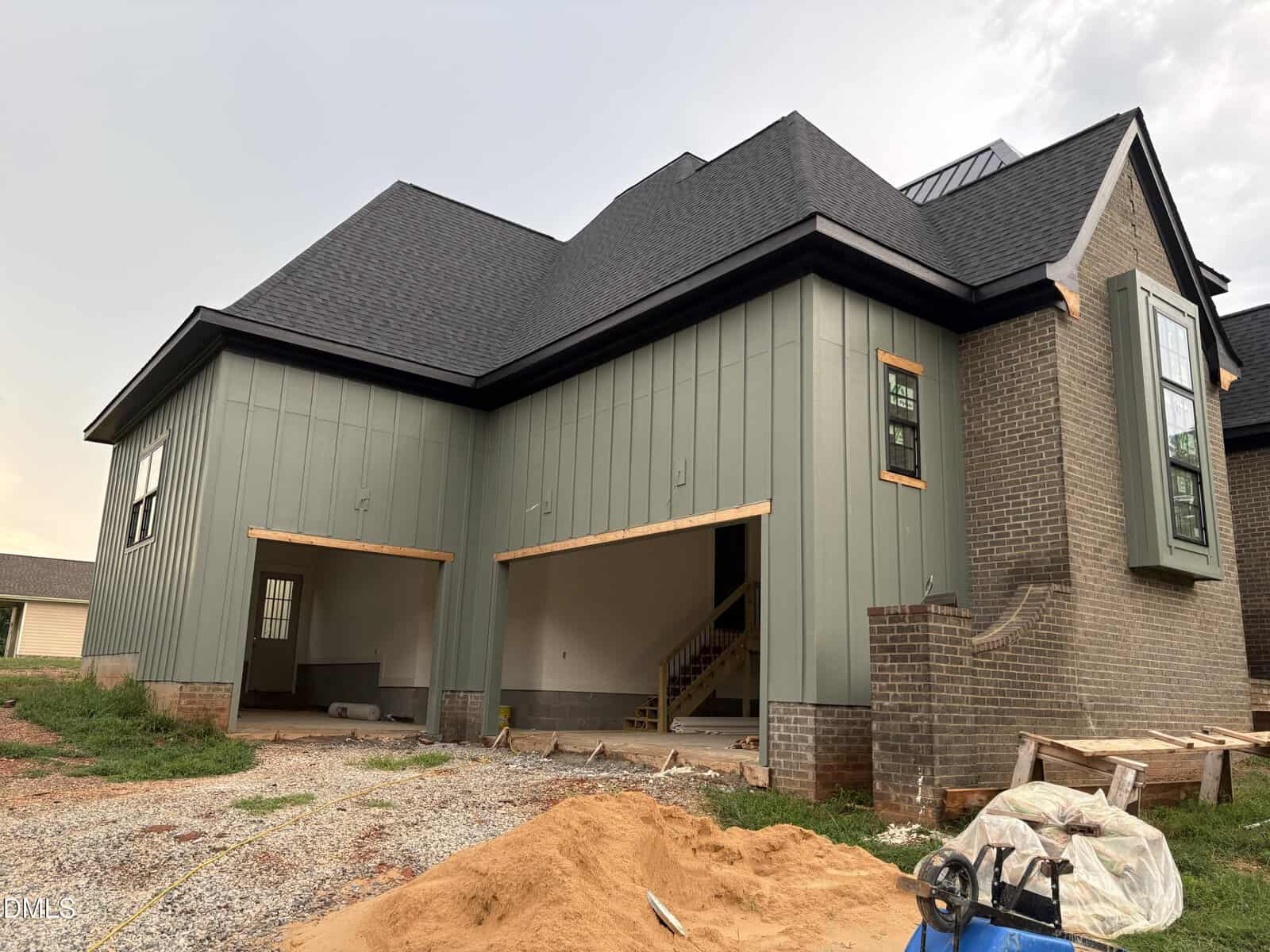 Partially constructed house with green siding, exposed garages, and unfinished landscaping. Tools, a blue barrel, wood, and construction materials are scattered in the foreground. The house has black shingles and large windows.