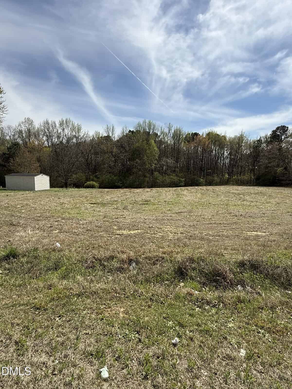 A grassy, open field with scattered debris in the foreground, bordered by a line of trees in the background. A small white shed is visible to the left, and the sky above is partly cloudy with a visible contrail.