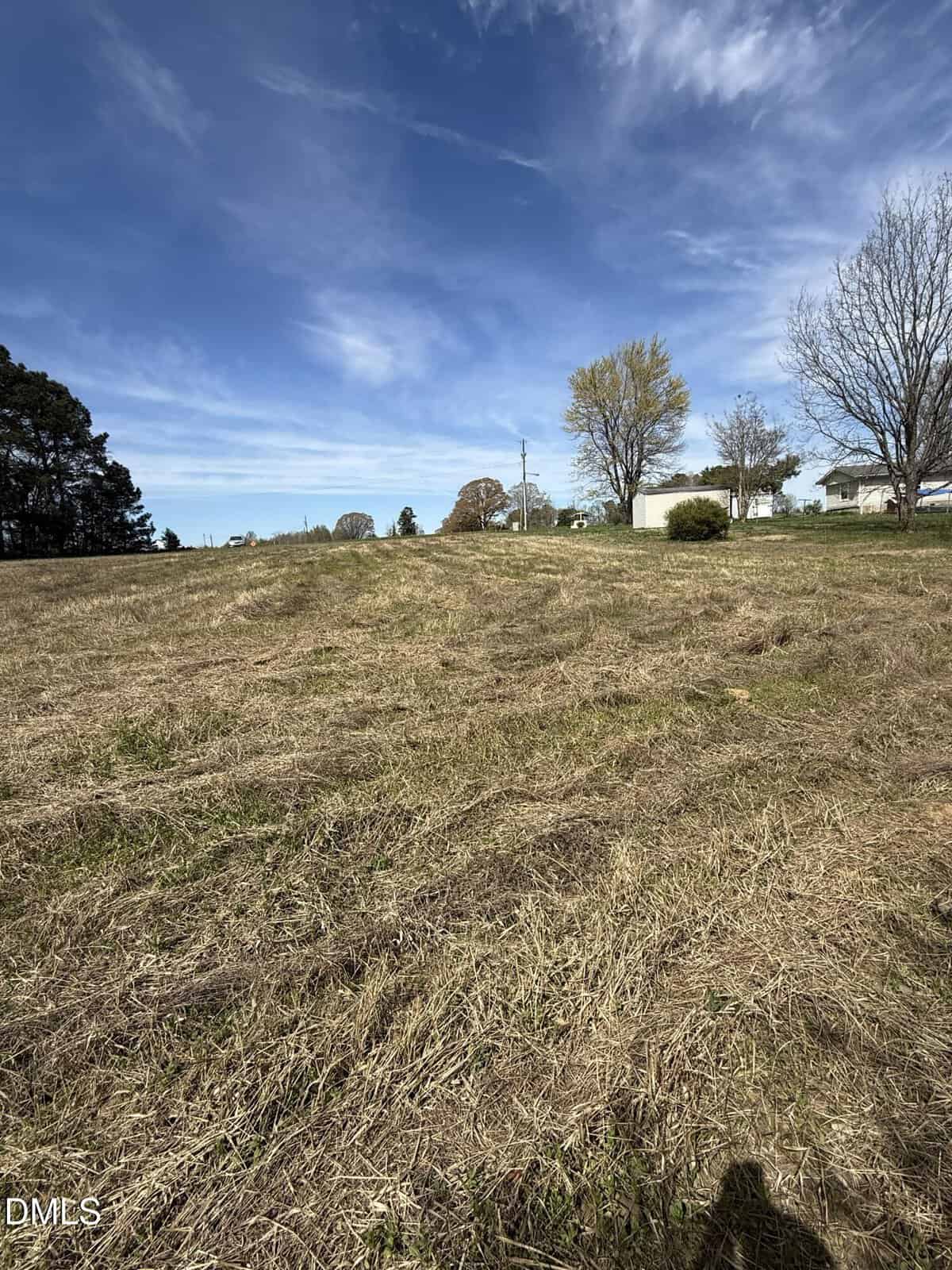 A dry, grassy field with a few trees and houses in the background under a blue sky with scattered clouds. The ground appears uneven and there is a shadow at the bottom edge of the image.