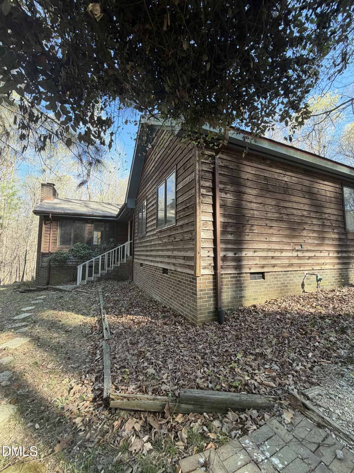 A rustic wooden house with brick foundation and sloped roof sits in a wooded area. A white handrail lines a ramp to the entrance. Fallen leaves cover the ground and some overhanging branches frame the top of the image.