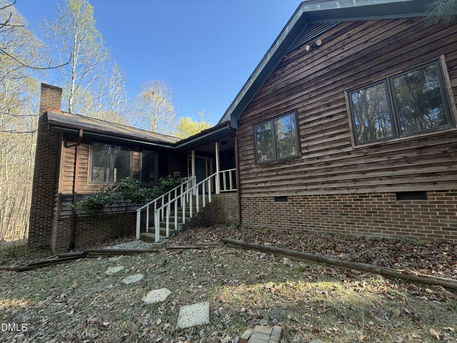 A wooden house with brick foundation, large windows, and a covered porch with white railing. The yard has patches of grass, scattered leaves, and stone steps leading to the entrance. Trees surround the home.