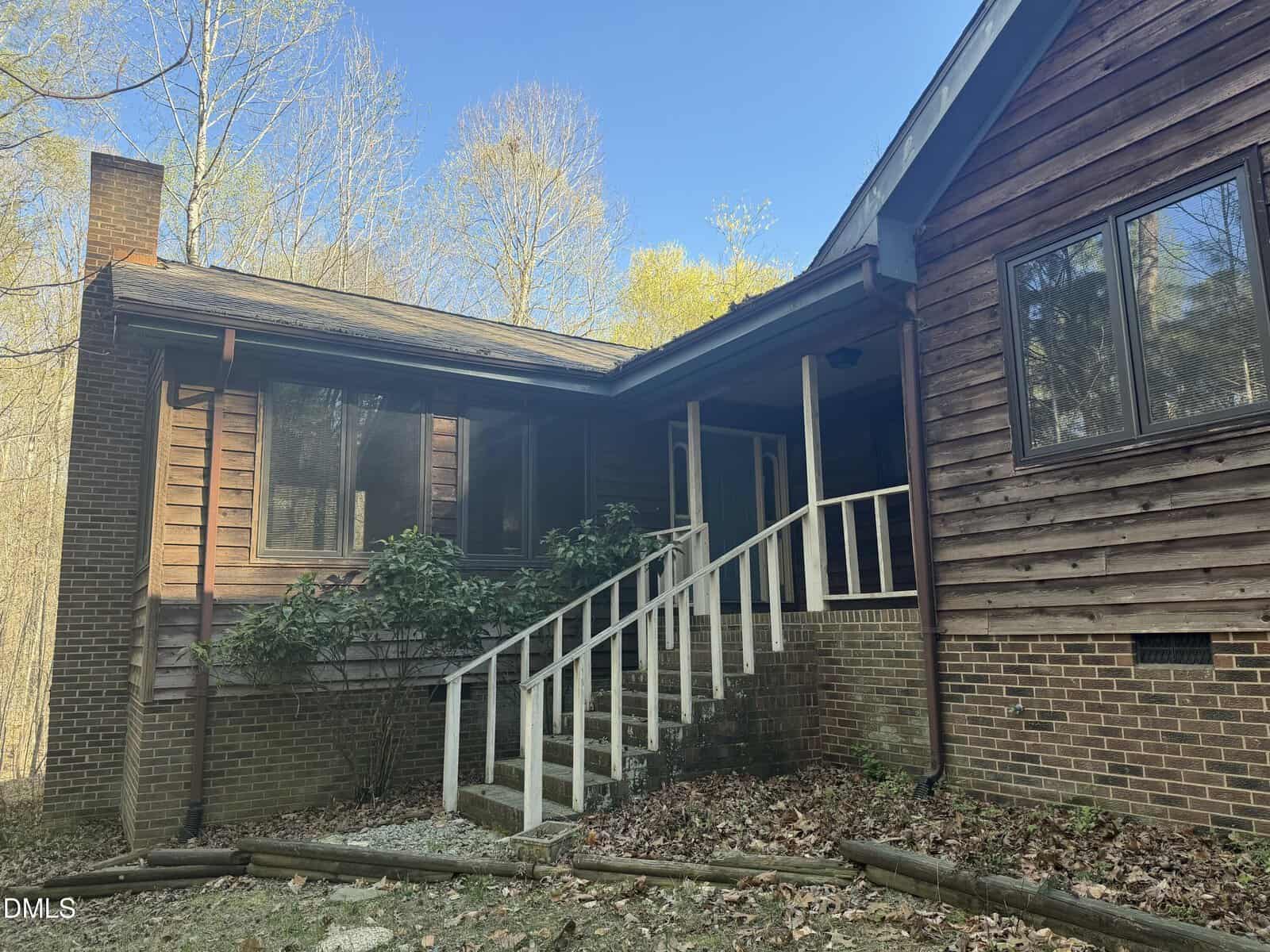 A rustic wooden house with a brick foundation, large windows, and a white railing leading up steps to the front porch. The yard is covered with leaves and surrounded by tall trees with bare branches.