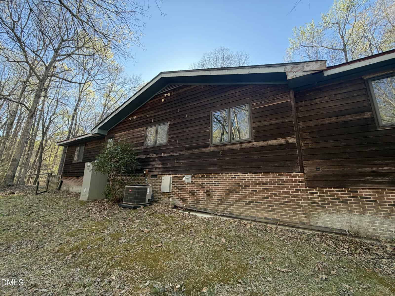 Single-story house with a brick foundation and dark wooden siding. There are three windows, an outdoor air conditioning unit, and leafless trees in the background. The lawn is covered with dry leaves.