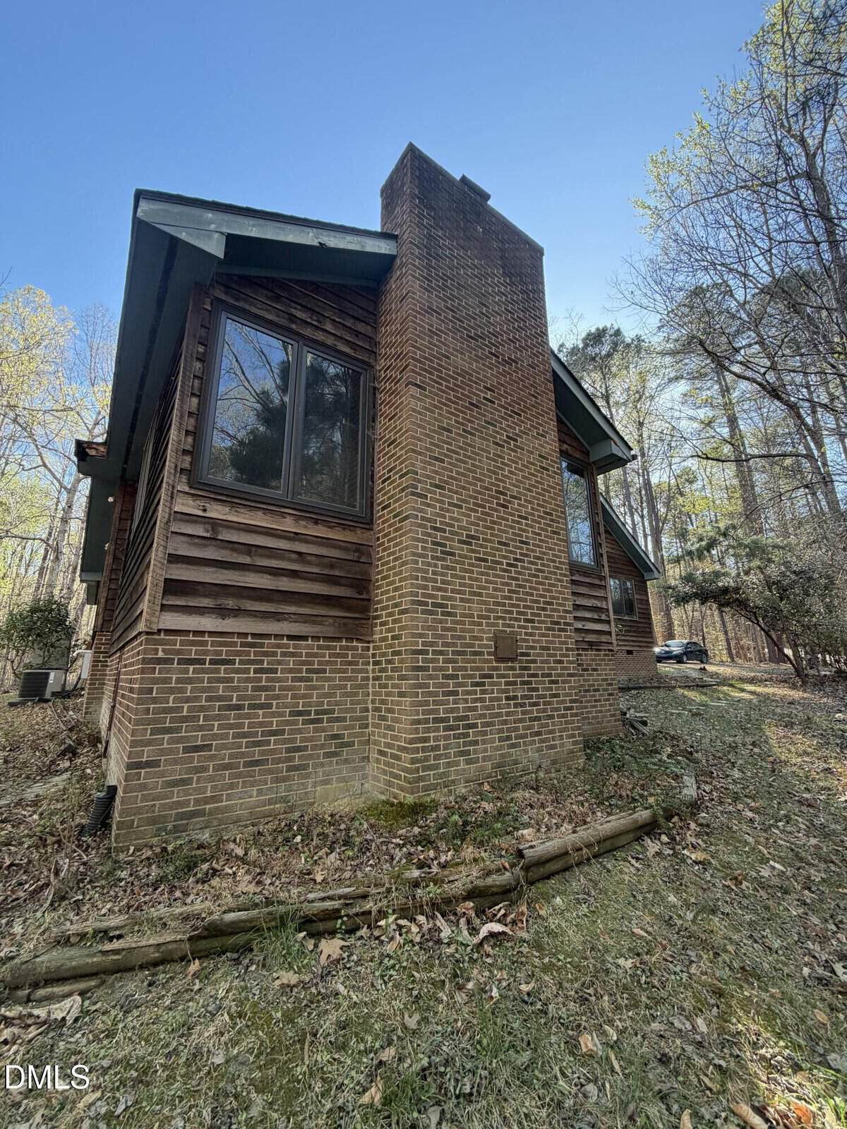 A two-story house with a prominent brick chimney and wood siding, surrounded by trees and a grassy yard with scattered leaves; a car is partially visible in the background.