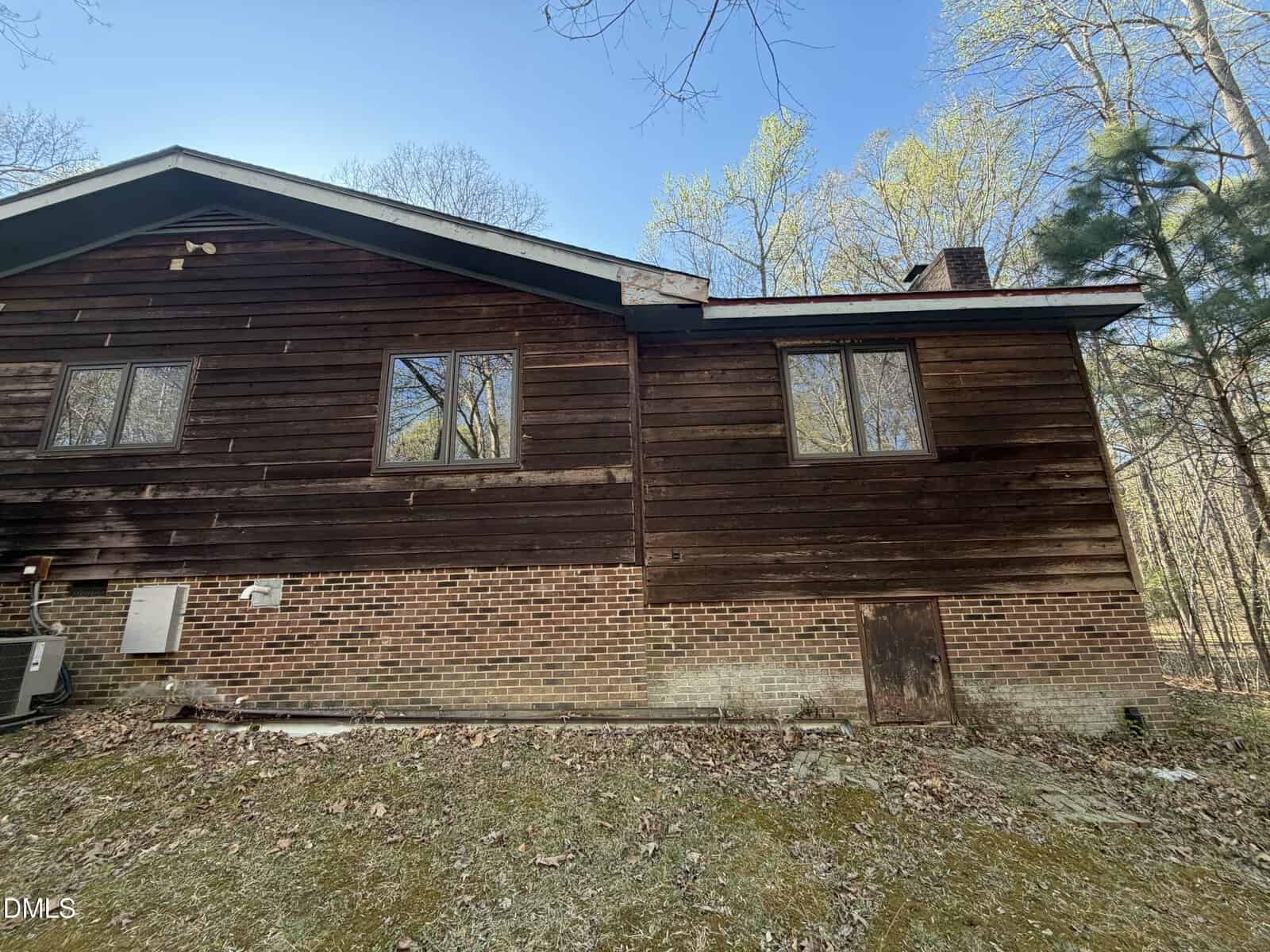 The image shows the side of a house with a brick foundation and dark wood siding. There are three windows, an exterior air conditioning unit, and a small door at ground level. Trees and grass are visible in the background.