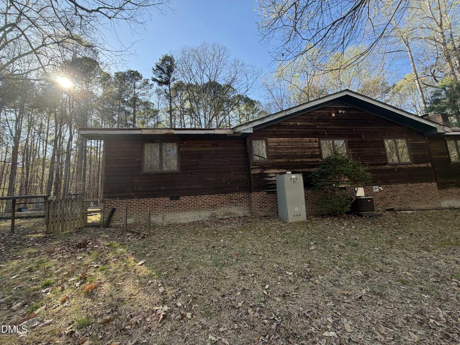 A single-story house with dark wood siding and brick foundation stands in a wooded yard. Sunlight filters through tall trees. An air conditioning unit and utility box are next to the house. Dry leaves cover the ground.