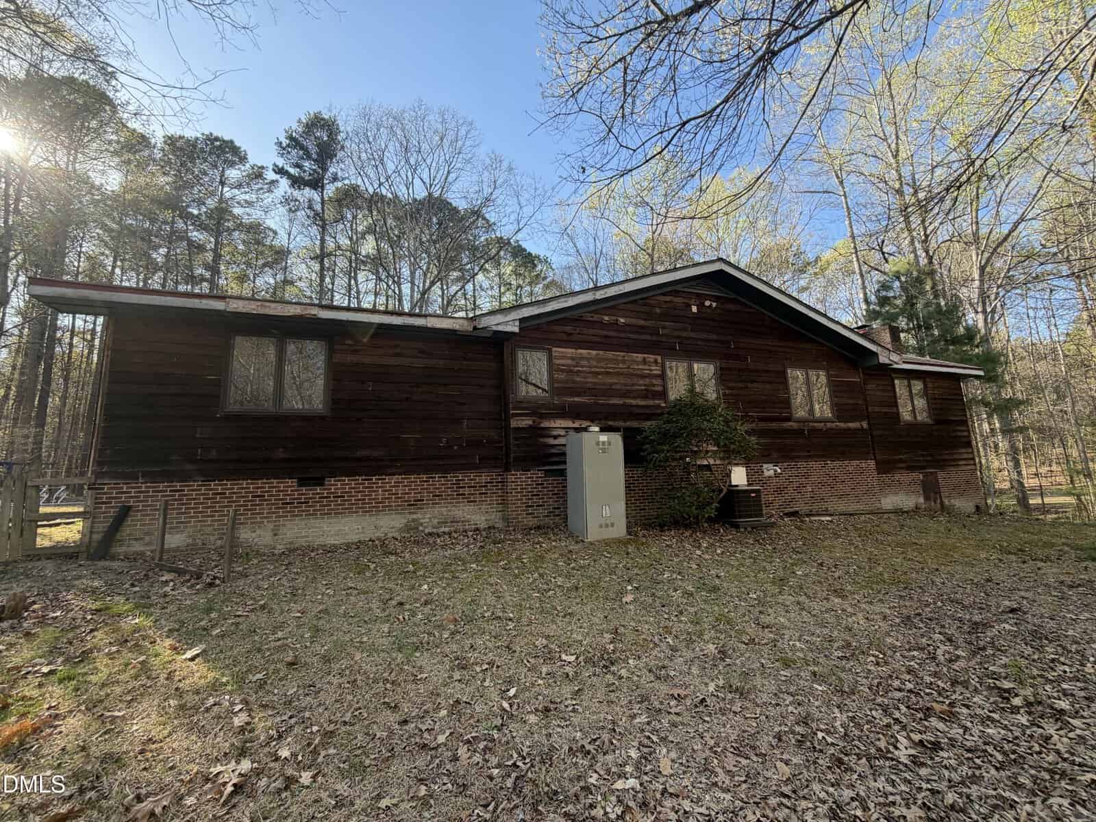 A single-story wooden and brick house with several windows, surrounded by trees. An electrical box and bushes are near the wall. The yard is covered with fallen leaves and grass. The sky is clear and sunny.