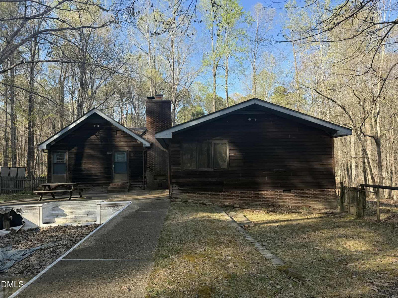 A brown, single-story wooden house with a brick foundation, two peaked roof sections, a chimney, and a small patio area, surrounded by trees and a grassy yard.
