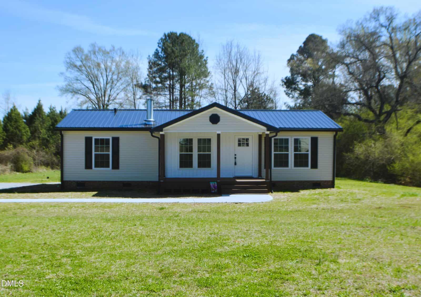Single-story white house with dark trim and a blue metal roof, set on a large grassy yard with trees in the background on a sunny day, located at 1159 No Bottom Road in Warrenton.