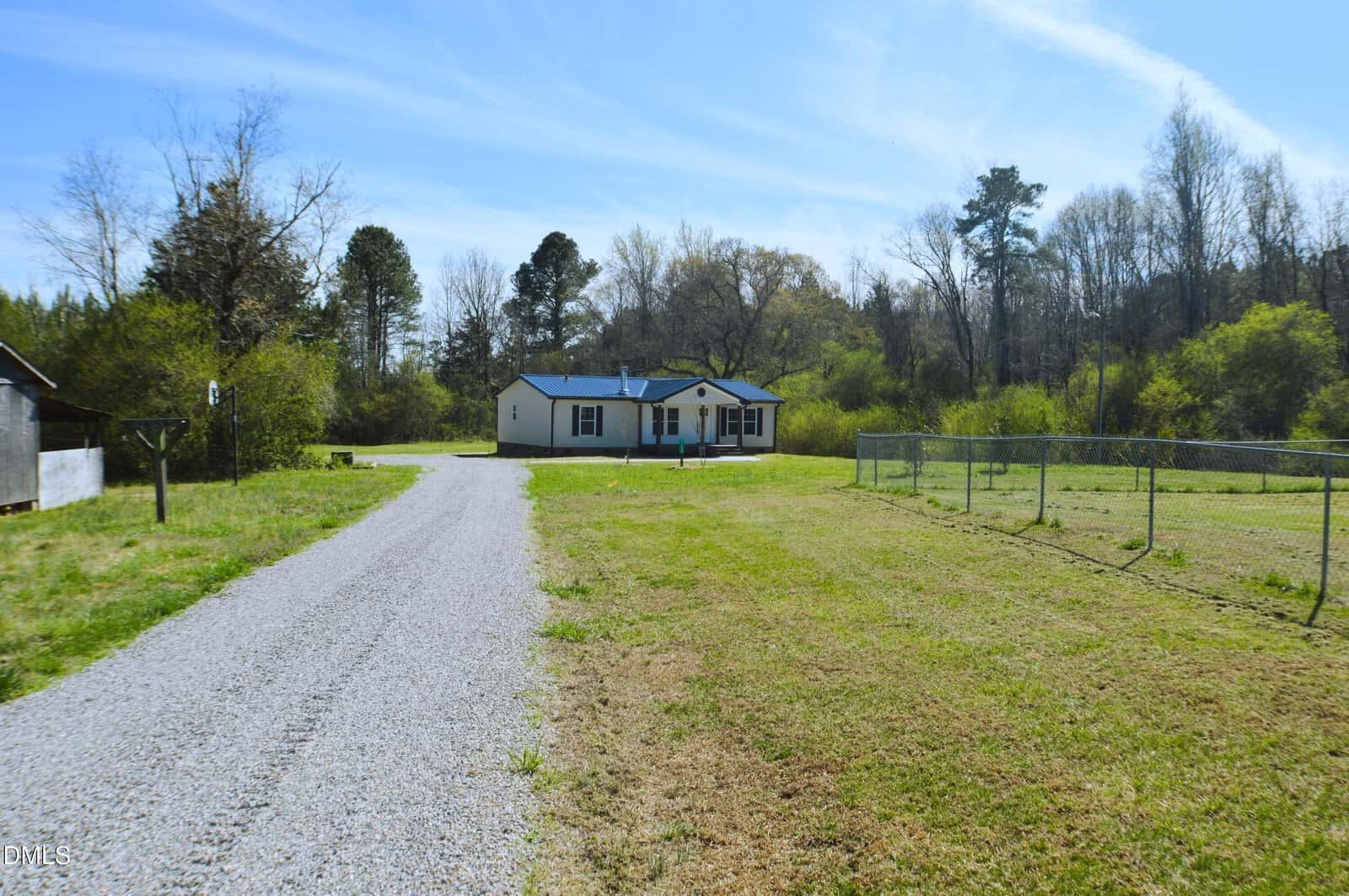 A gravel driveway leads to a single-story yellow house with a blue roof, surrounded by grass, trees, and a wire fence under a clear blue sky.