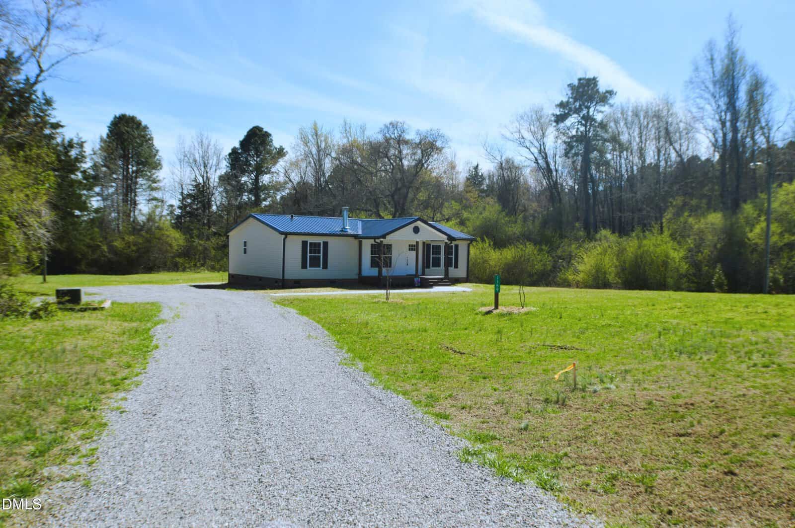 A single-story white house with a dark roof sits at the end of a gravel driveway, surrounded by grass, trees, and a partly cloudy blue sky.