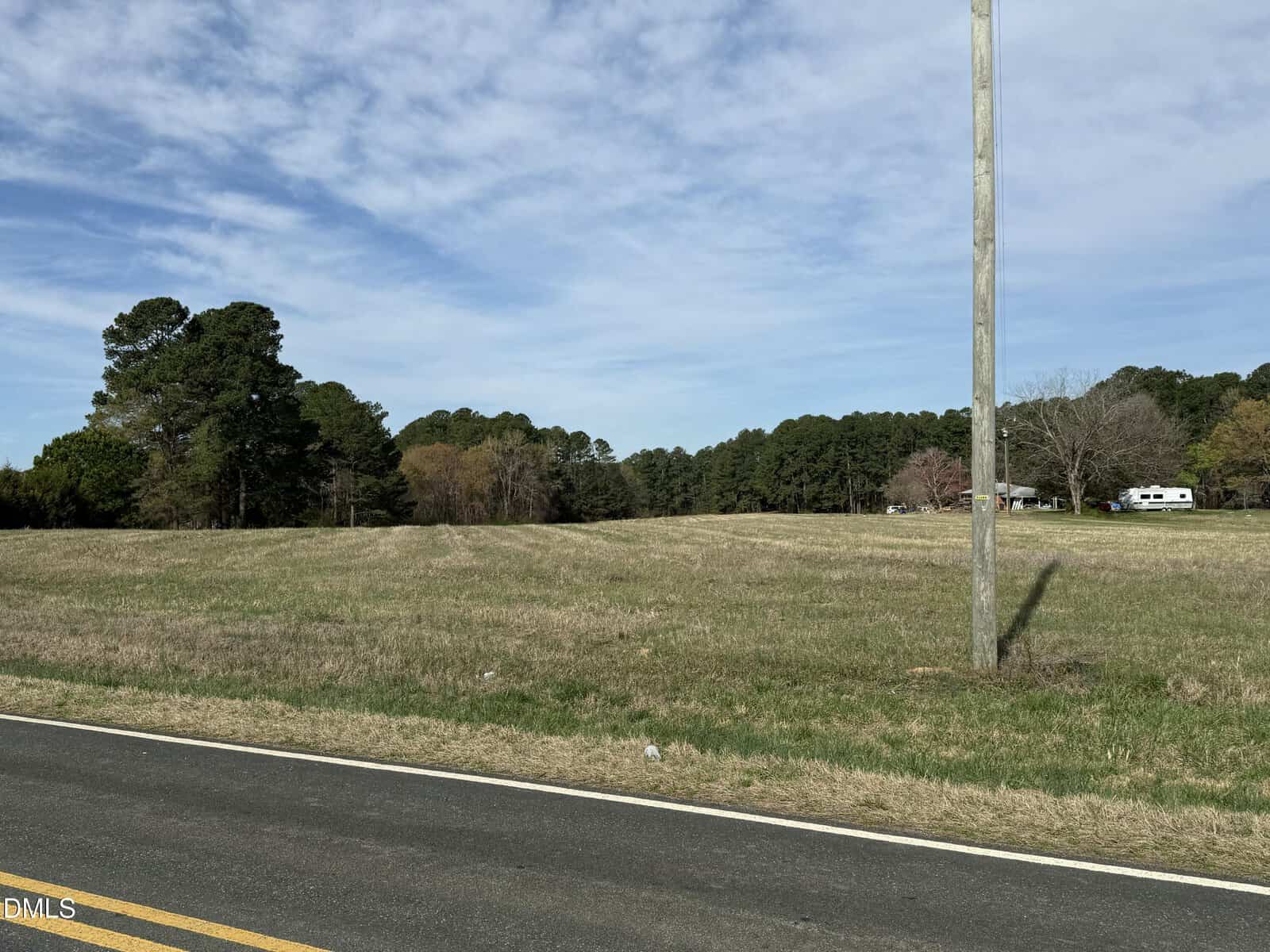 A grassy open field bordered by trees under a partly cloudy sky, with a paved road and a utility pole in the foreground. A white vehicle is visible in the distance near some trees.