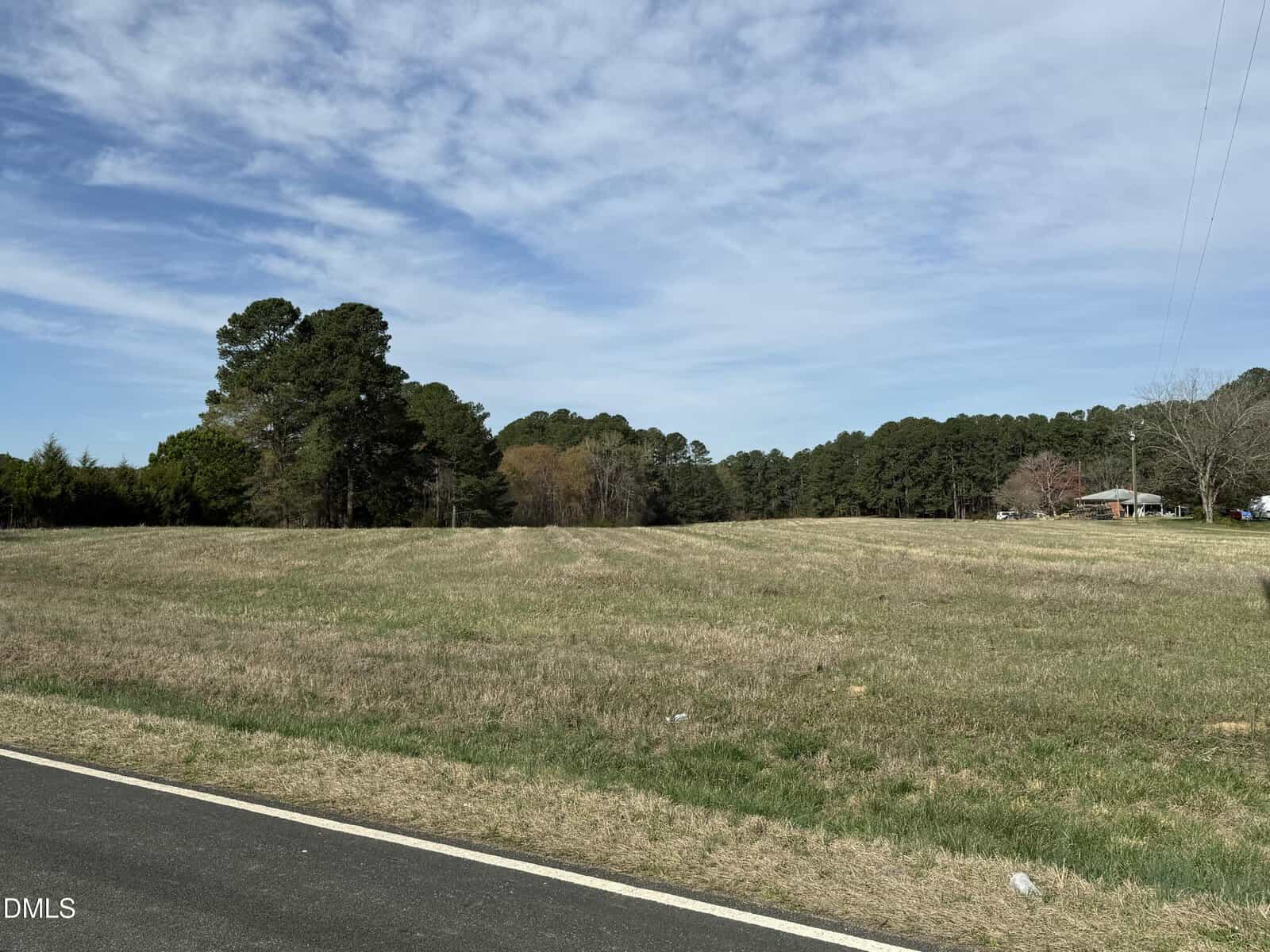 A grassy field bordered by trees under a partly cloudy sky, with a paved road in the foreground and a house visible in the distance on the right.