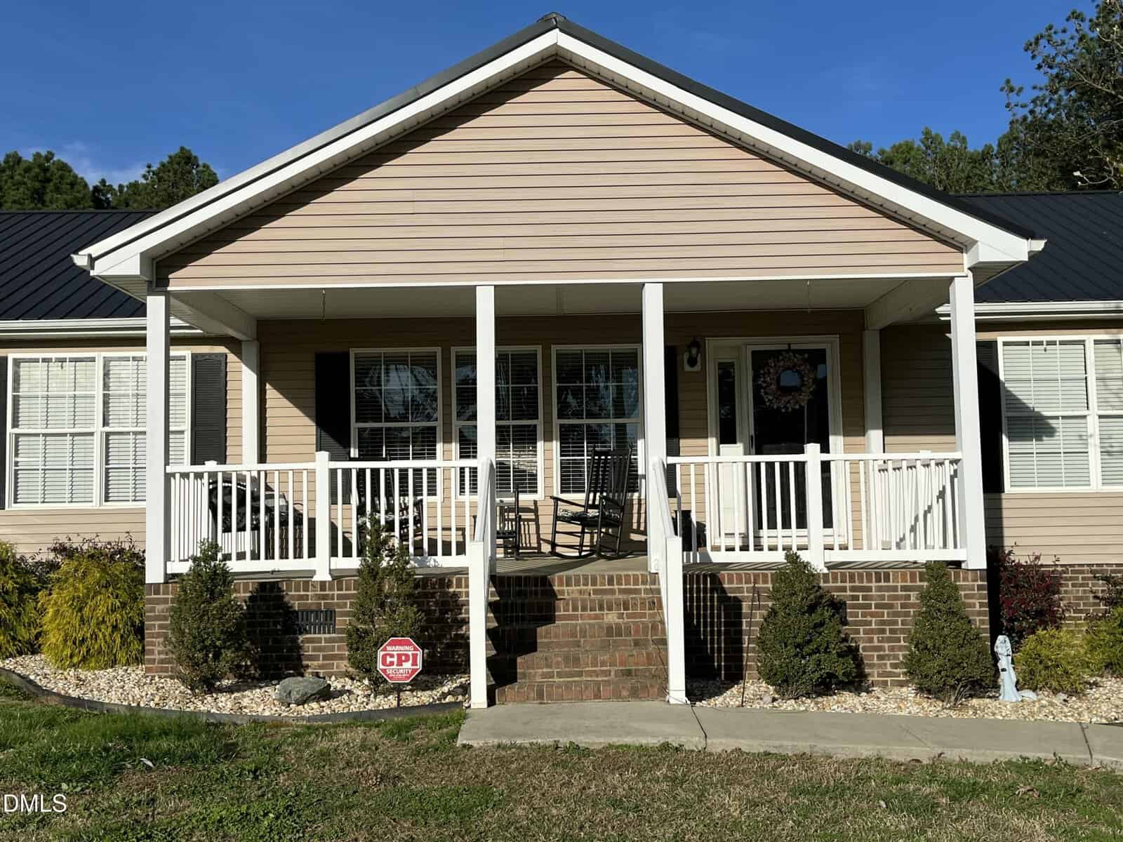 Single-story tan house with white trim, a covered front porch, brick steps, two rocking chairs, and small shrubs in front. A security sign is placed in the landscaping to the left of the steps.