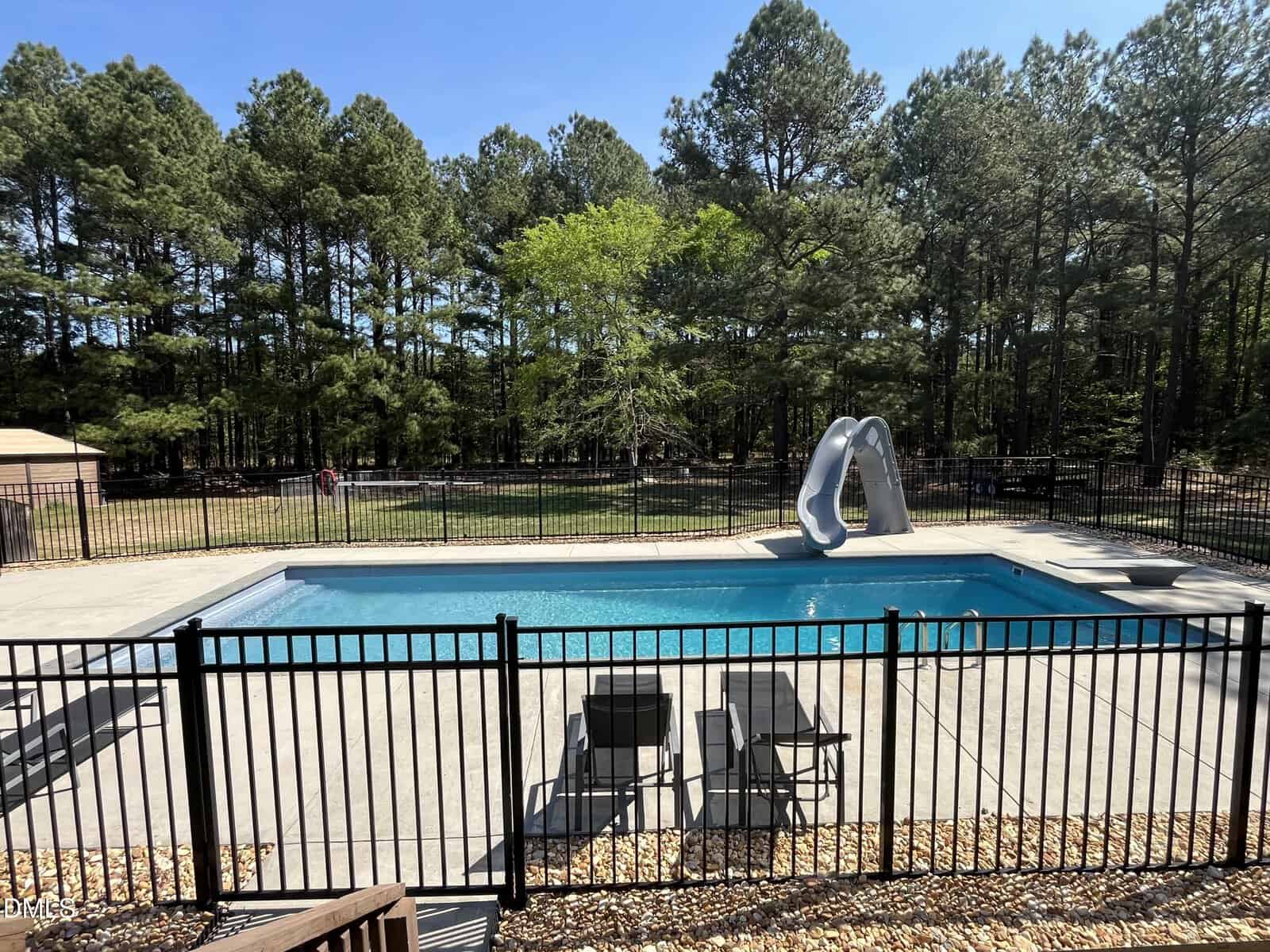 A fenced-in outdoor pool with a slide, surrounded by lounge chairs and trees in the background on a sunny day.