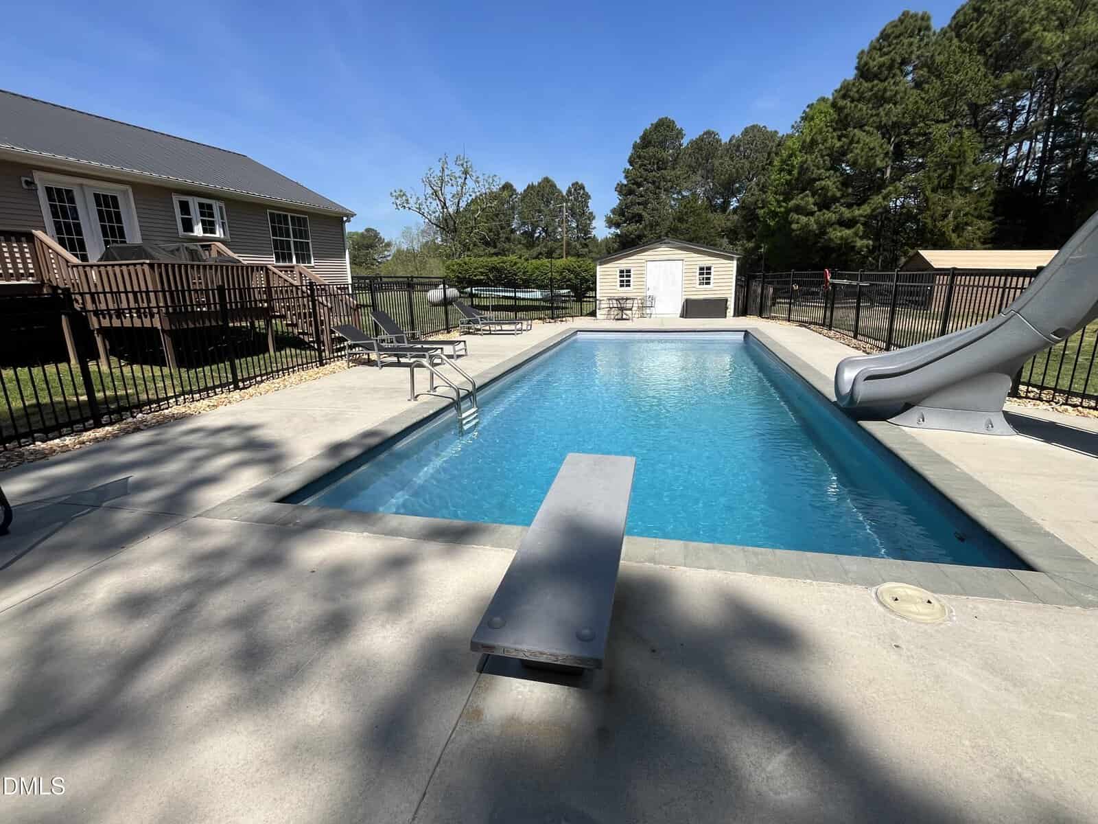 Rectangular outdoor swimming pool with a diving board at the near end and a slide at the far end, surrounded by a concrete deck, black fence, and trees. A house and shed are visible in the background.