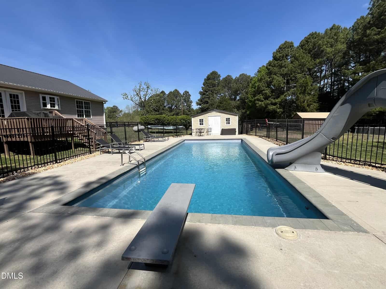 A backyard swimming pool with a diving board and a slide, surrounded by a concrete deck and black metal fence. There is a house and a shed in the background, with trees and clear blue sky.