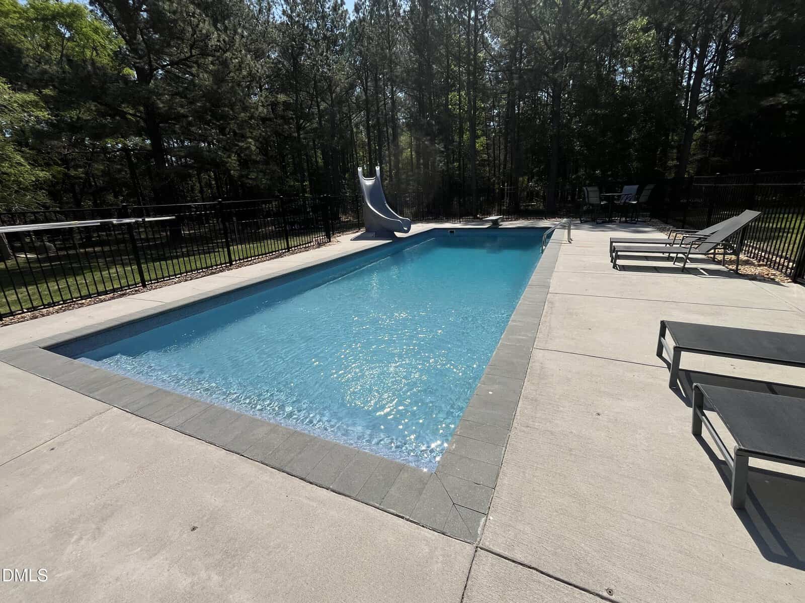 Rectangular outdoor swimming pool with clear blue water, bordered by gray tiles and concrete patio; a blue slide at the far end and lounge chairs on the right, surrounded by a black metal fence and trees in the background.