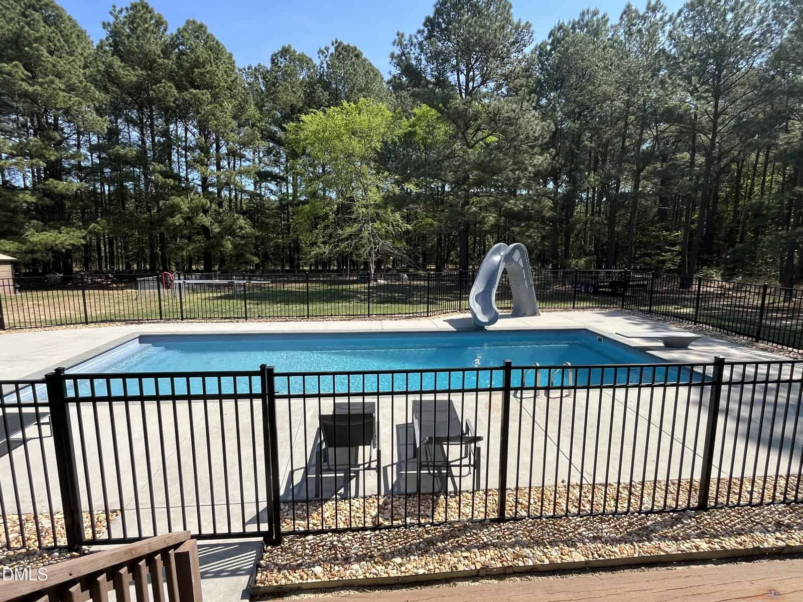 A fenced outdoor swimming pool with a slide, surrounded by lounge chairs and trees in the background on a sunny day.